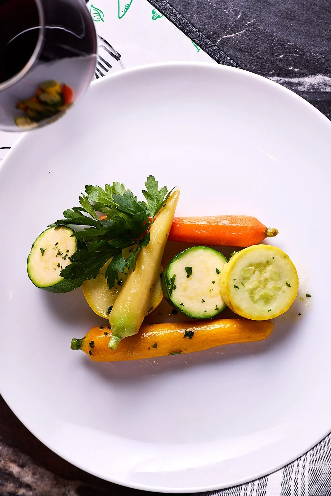 Plate with sliced zucchini, yellow squash, carrots, cucumber, and parsley, with a glass of red wine in the corner.