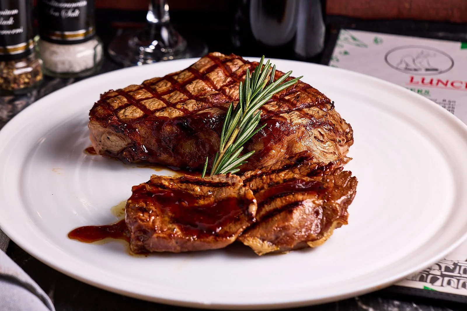A cooked steak with grill marks, garnished with a sprig of rosemary, served with sauce on a white plate.