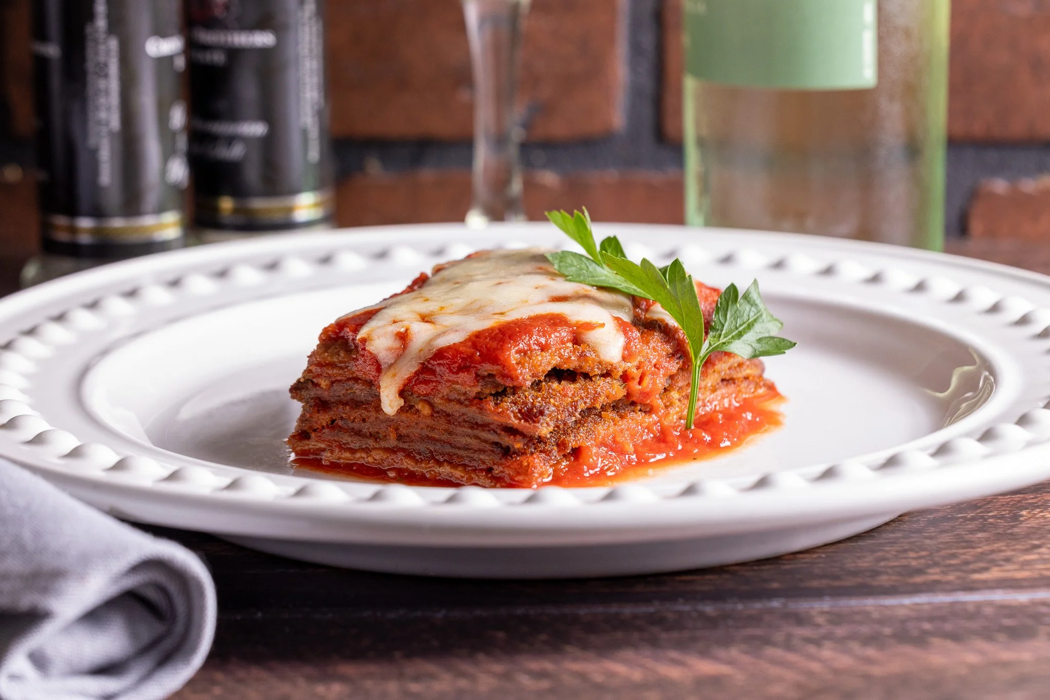 A serving of lasagna on a white decorative plate garnished with a sprig of parsley, with a brick wall and green glass in the background.
