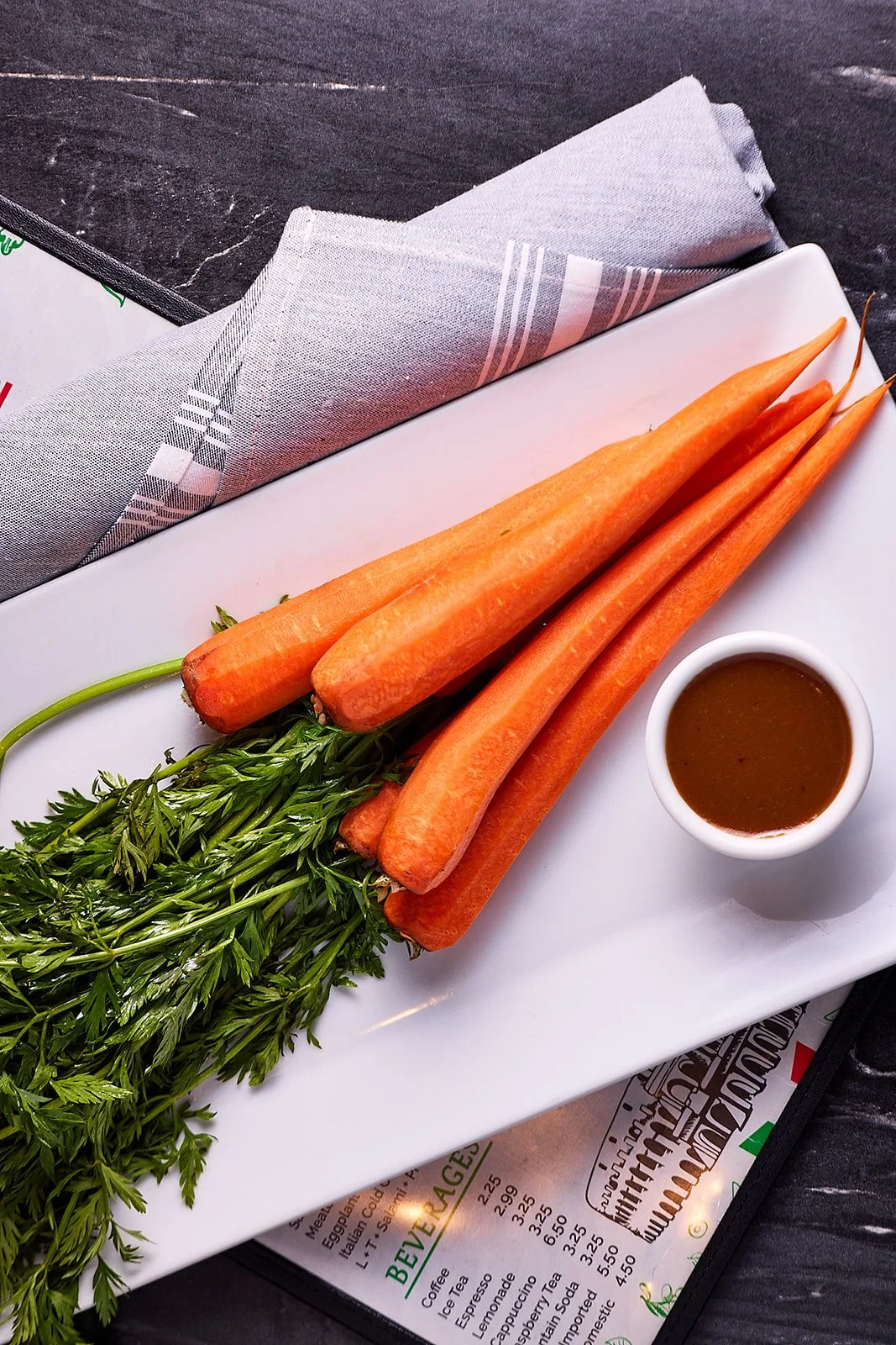 Fresh carrots with leafy greens and a small bowl of dipping sauce on a white rectangular plate.