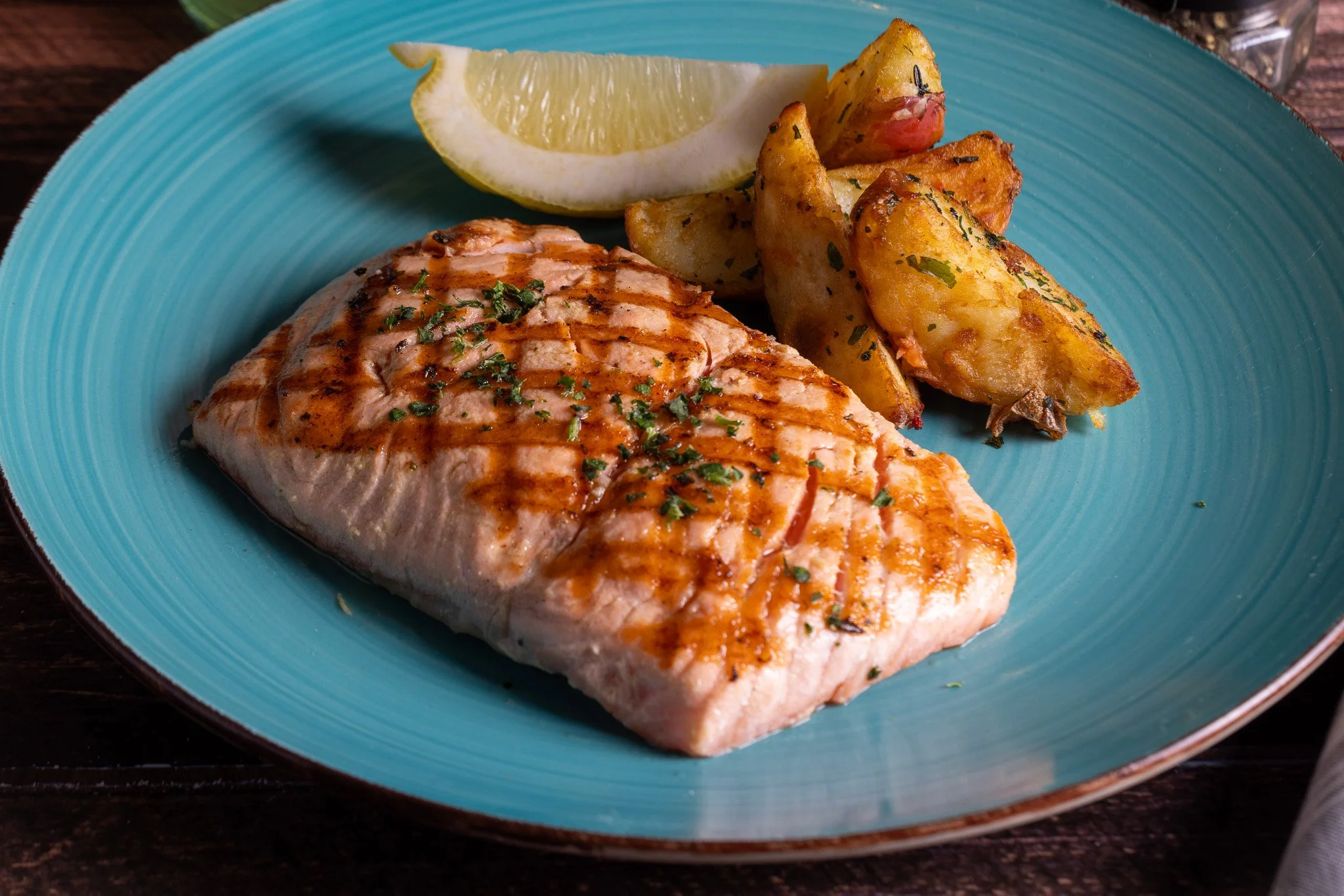 Grilled salmon filet with lemon wedge and roasted potato wedges on a turquoise plate.