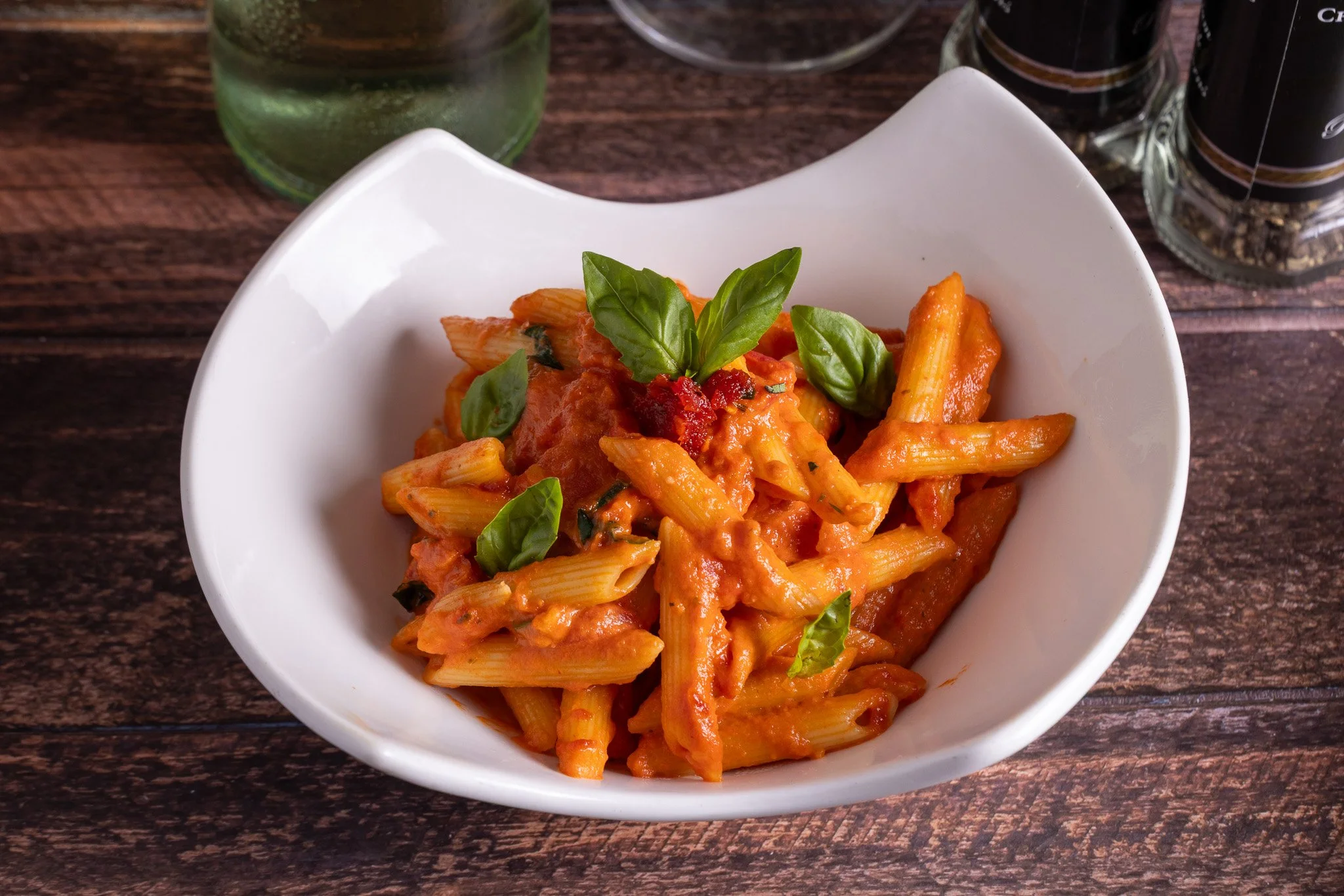 A white bowl filled with penne pasta in tomato sauce, garnished with fresh basil leaves, placed on a wooden table.