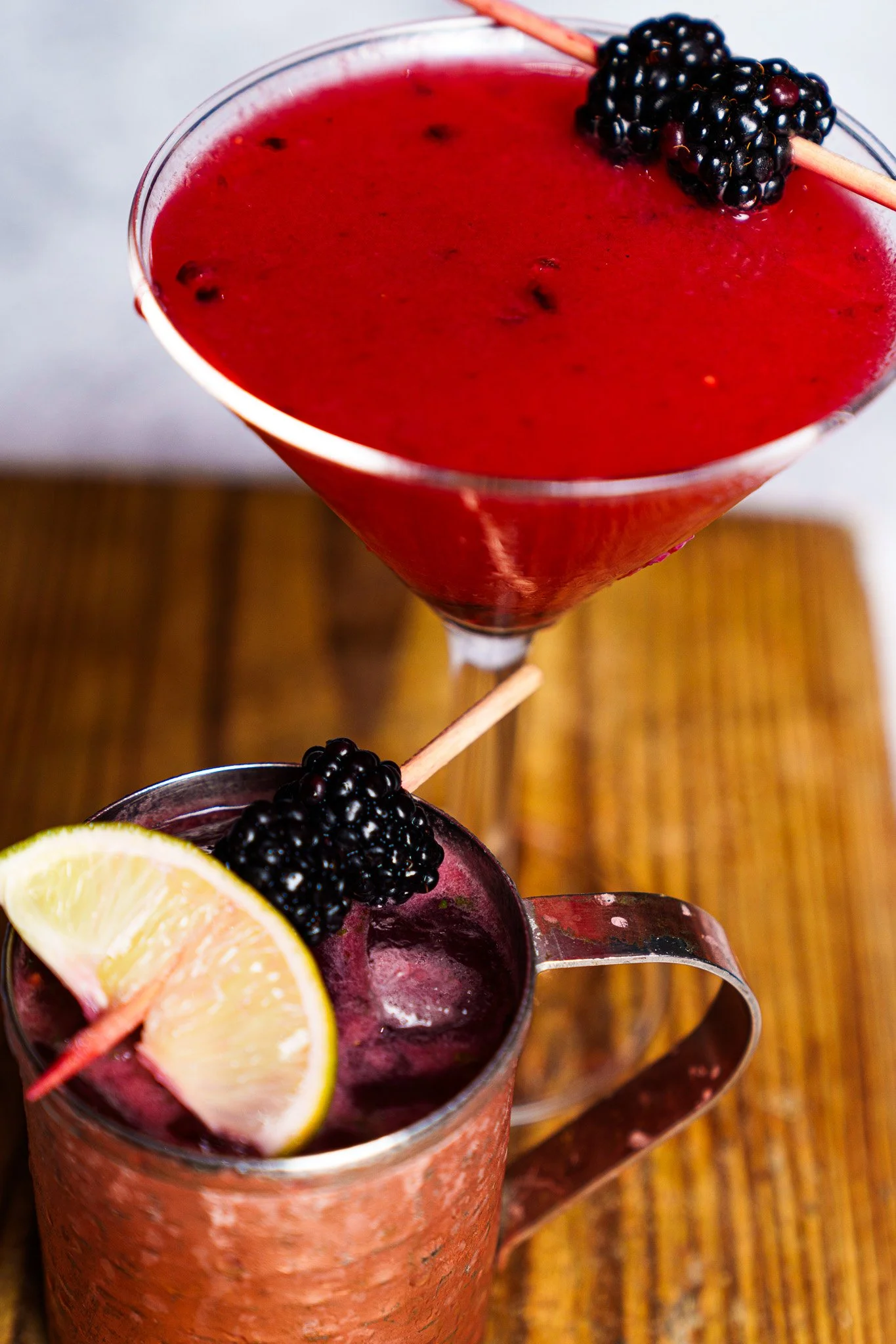 A close-up of two blackberry-infused cocktails; the top cocktail is in a martini glass with a blackberry skewer garnish, and the bottom cocktail is in a copper mug with a lemon wedge and blackberry skewer garnish, both placed on a wooden surface.