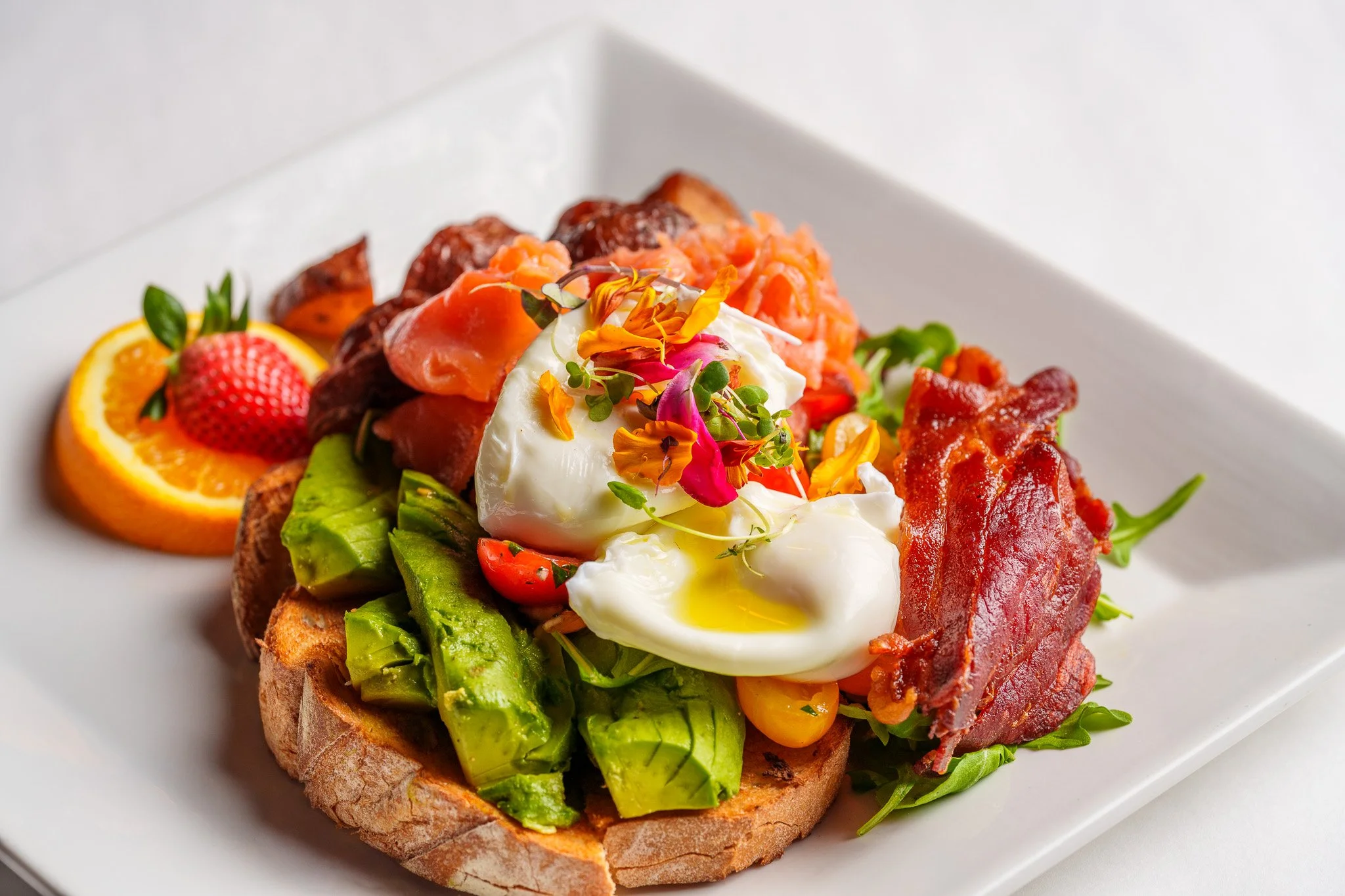 Open-faced avocado toast with poached egg, bacon, smoked salmon, fresh berries, orange slices, and edible flowers on a white plate.