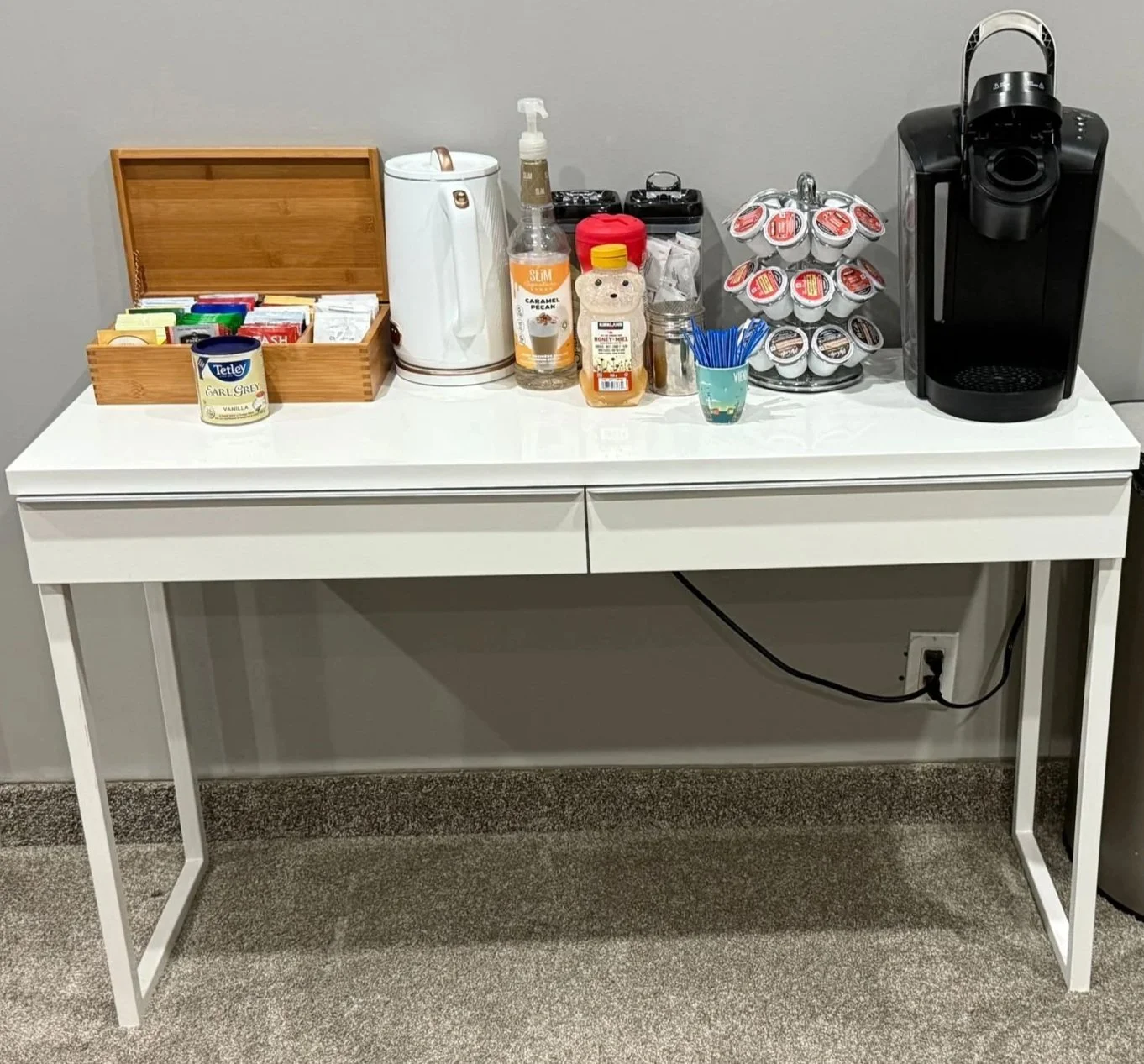 White coffee station with a Keurig coffee maker, coffee pods, syrup bottles, tea boxes, and a wooden box for tea bags.