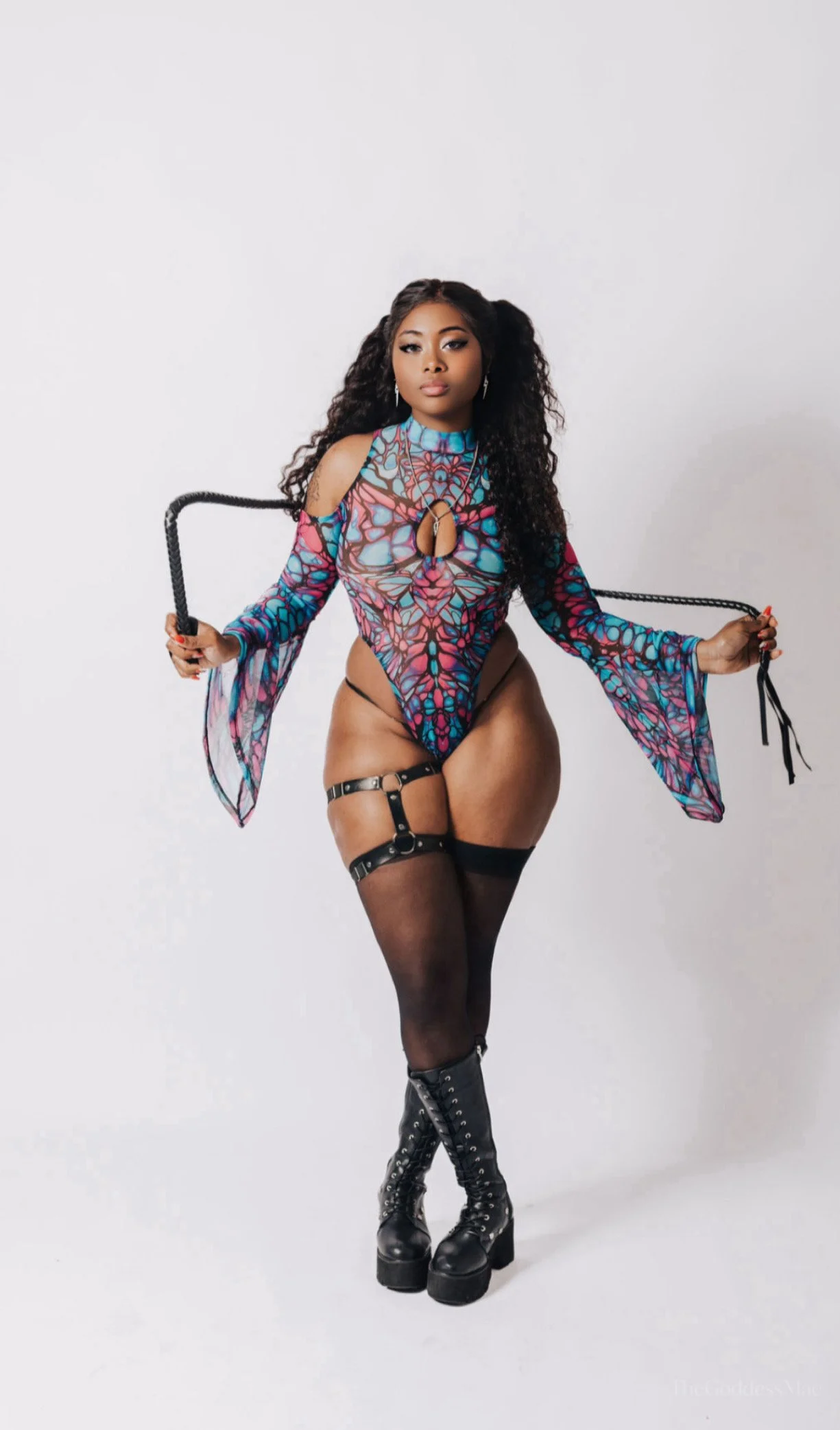 A woman with long curly hair wearing a colorful butterfly-themed bodysuit, black thigh-high stockings, and lace-up platform boots, standing against a plain white background and holding the ends of a black whip.