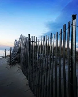 Beach with a wooden fence separating sand from dunes, under a partly cloudy sky.