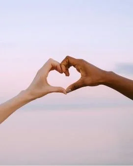 Two hands, one light-skinned and one dark-skinned, forming a heart shape against a pastel sky background.