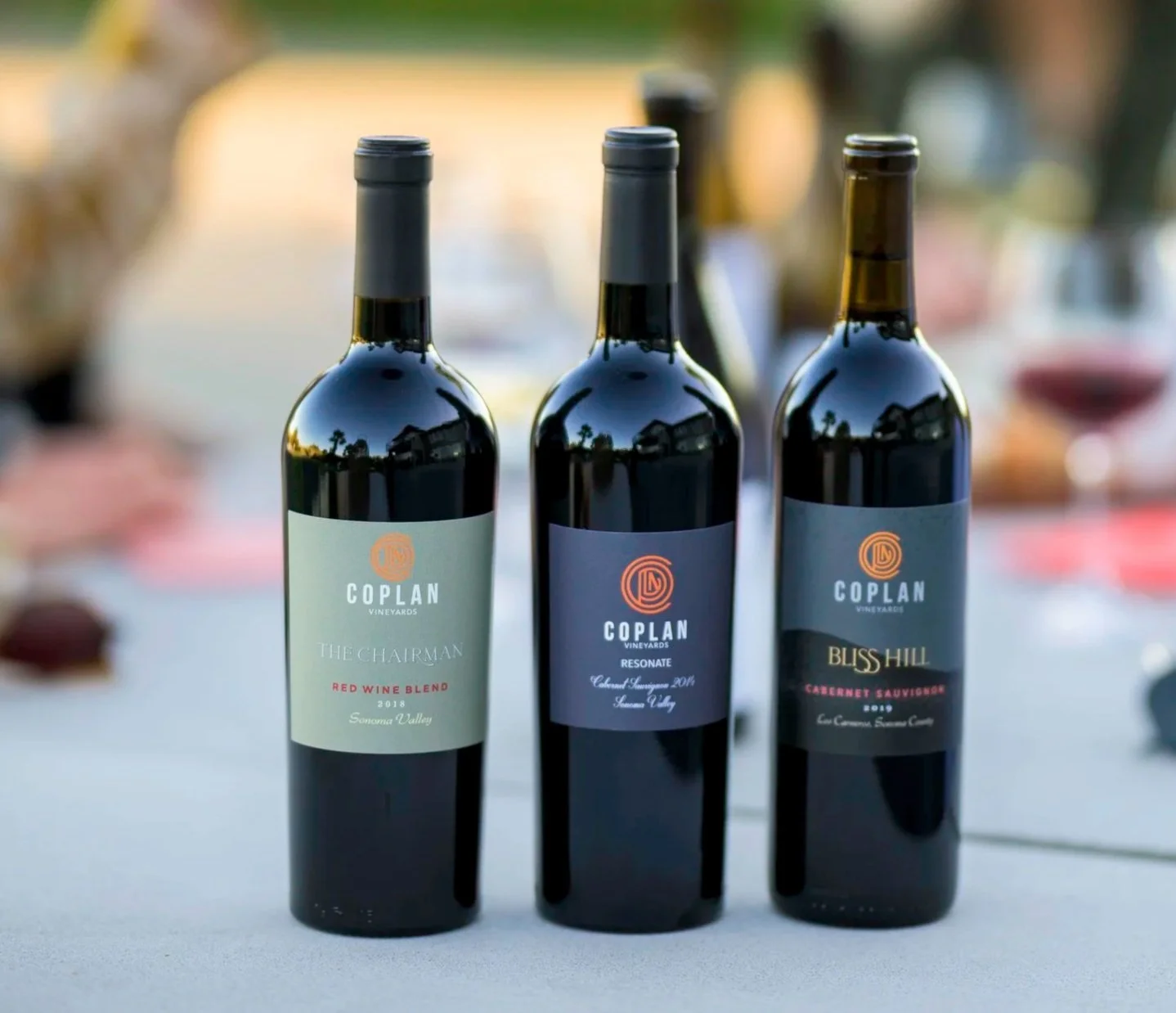Three bottles of red wine from Coplan Vineyards on a table at an outdoor event, with blurred background.