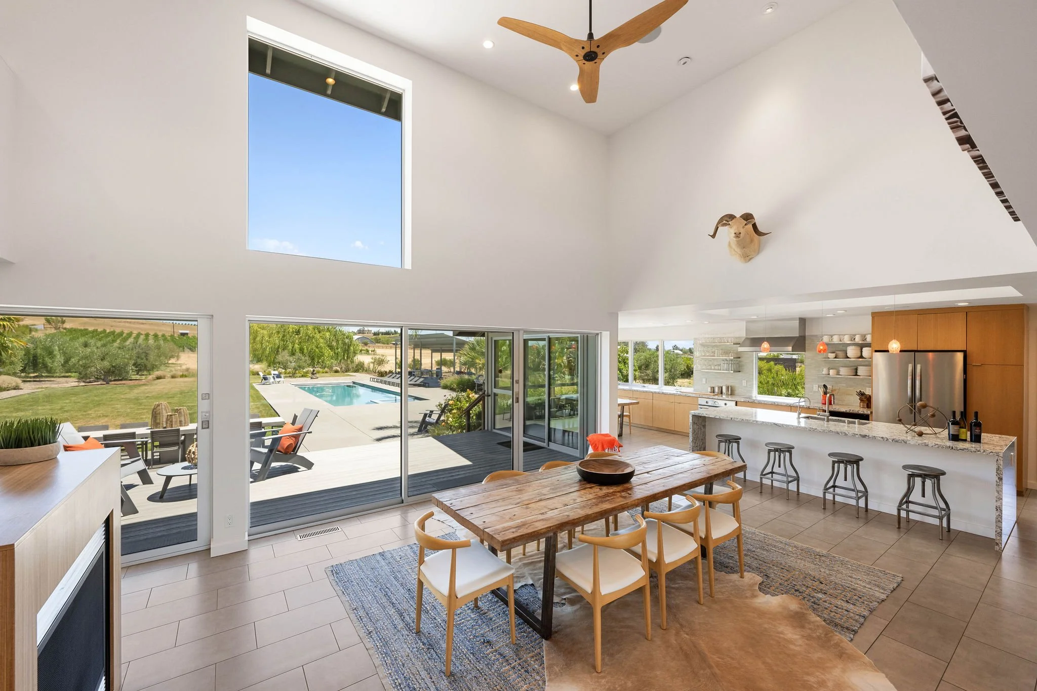 Bright modern open-concept dining and kitchen area with large glass sliding doors leading to an outdoor pool and yard, white walls, high ceilings, minimalist decor, and a mounted animal skull on the wall.