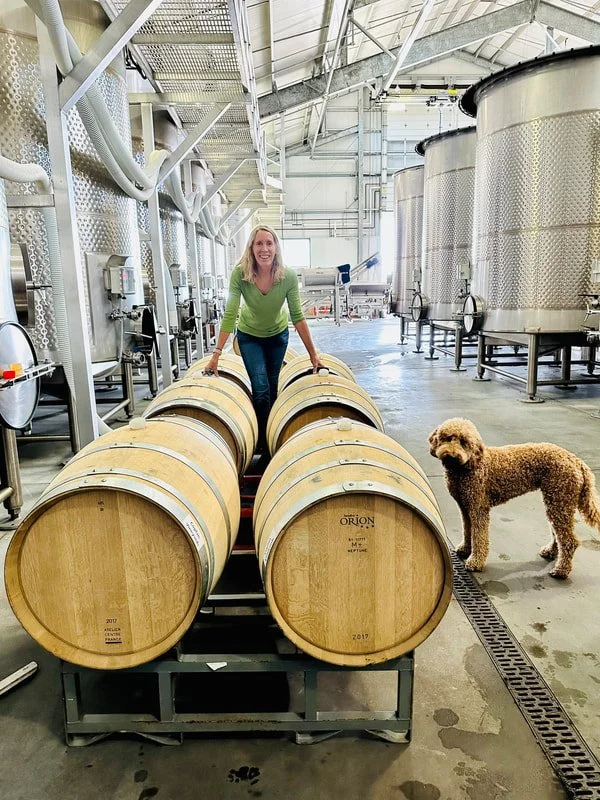 Jacqueline Yoakum, Coplan Vineyards, winemaker, with a dog standing on a wine barrel rack inside a winery with large stainless steel fermentation tanks.