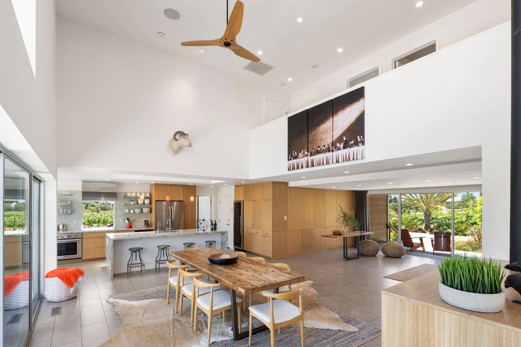 Open-concept modern kitchen and living area with wooden accents, a large dining table, and sliding glass doors to an outdoor patio with greenery, a palm tree, and outdoor seating. Ceiling fan and mounted animal head decor.