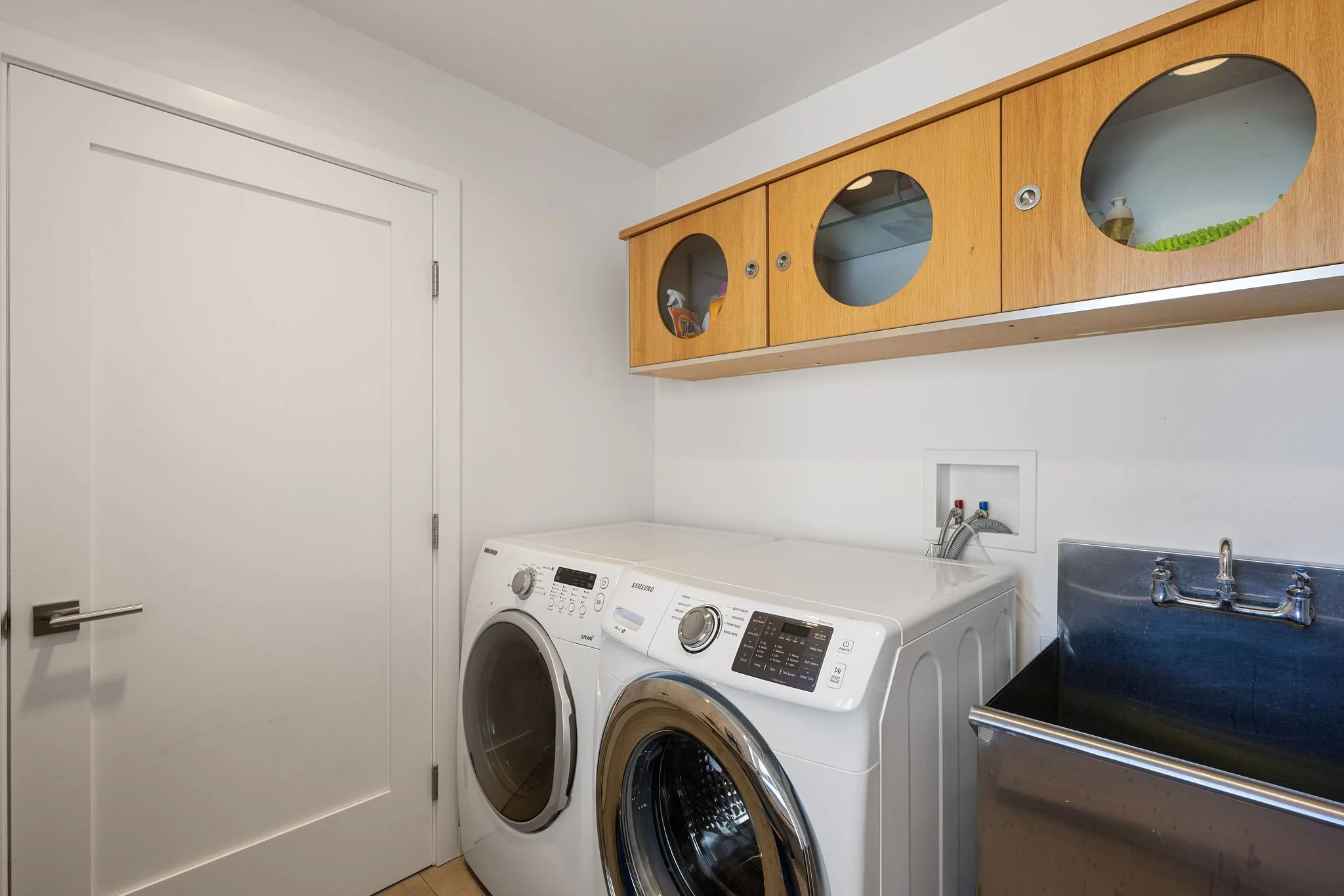 Laundry room with a white washer and dryer, a wooden wall cabinet with four oval windows, a utility sink, and a closed white door.