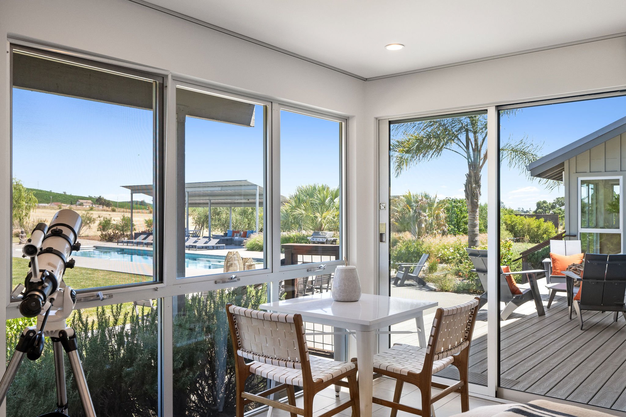View from inside a room with large glass windows looking out onto a backyard with a pool, outdoor seating, and palm trees.