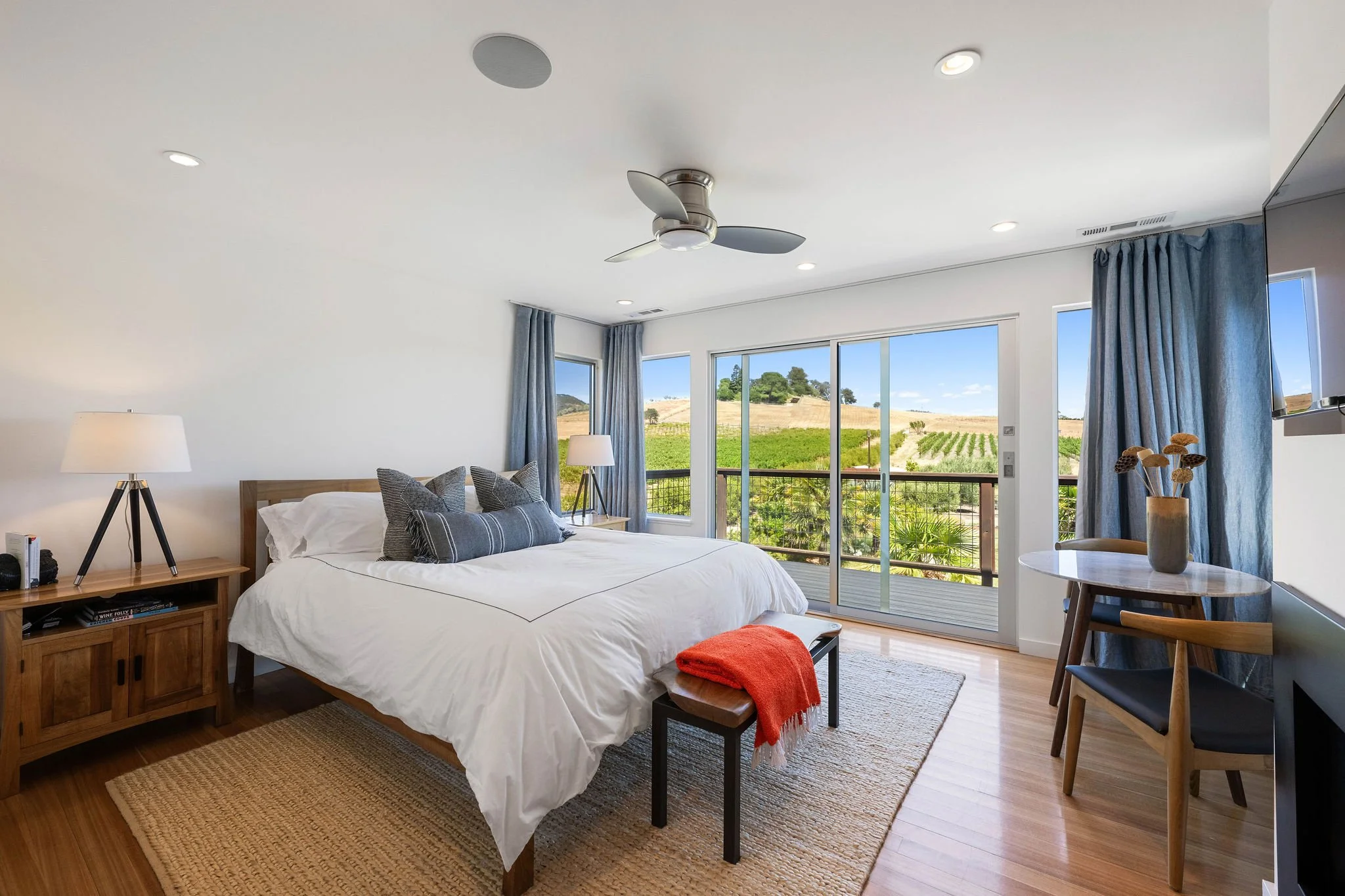 Bright bedroom with large sliding glass doors leading to a balcony, overlooking a vineyard landscape; white bed with decorative pillows, wooden nightstand with lamp, small table with a vase of flowers, blue curtains, ceiling fan, hardwood floors, and