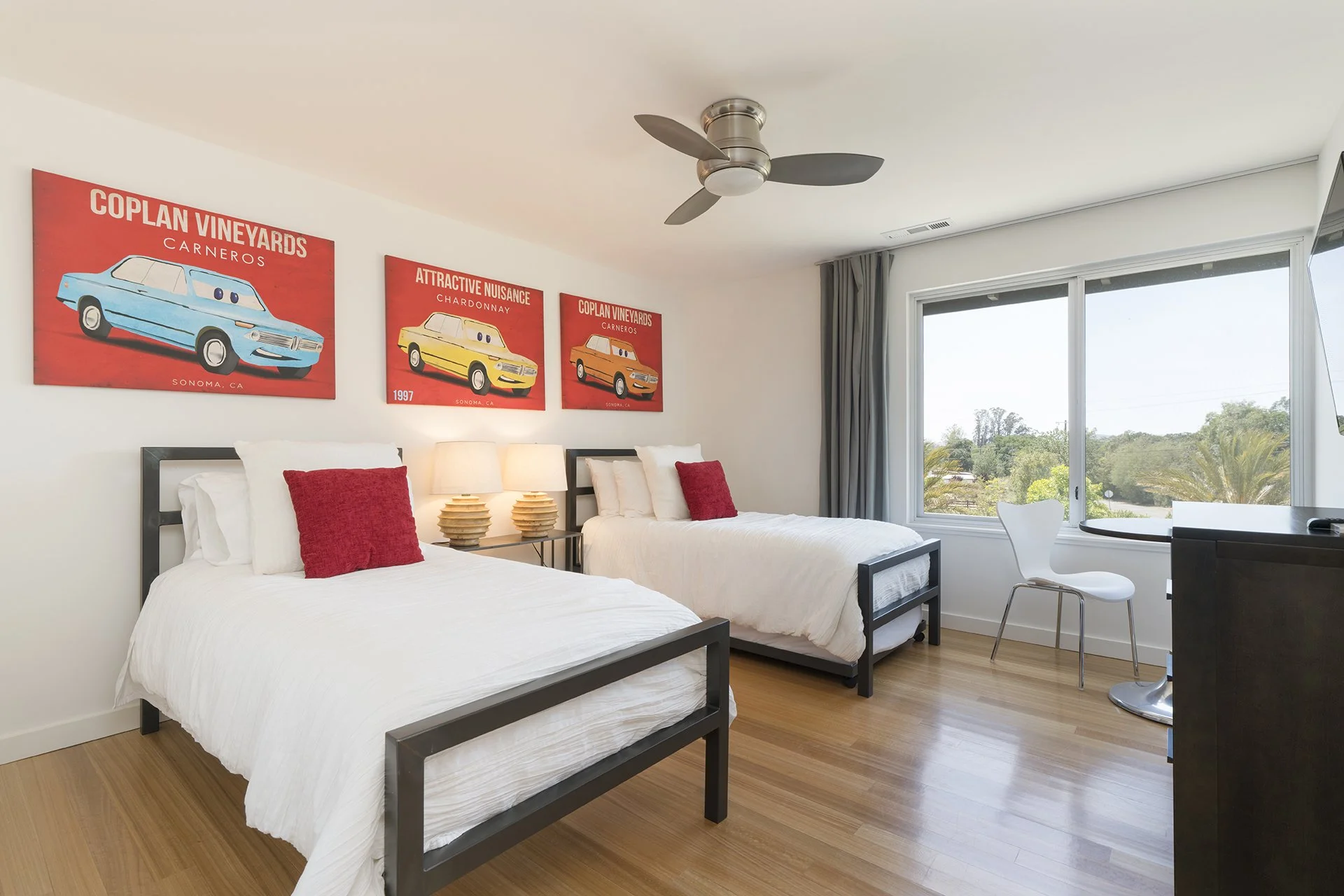 A hotel room with two beds, each with white bedding and red pillows. There is a nightstand with two lamps and three comic-style posters of cars on the wall. The room has a large window showing a view of trees and blue sky.