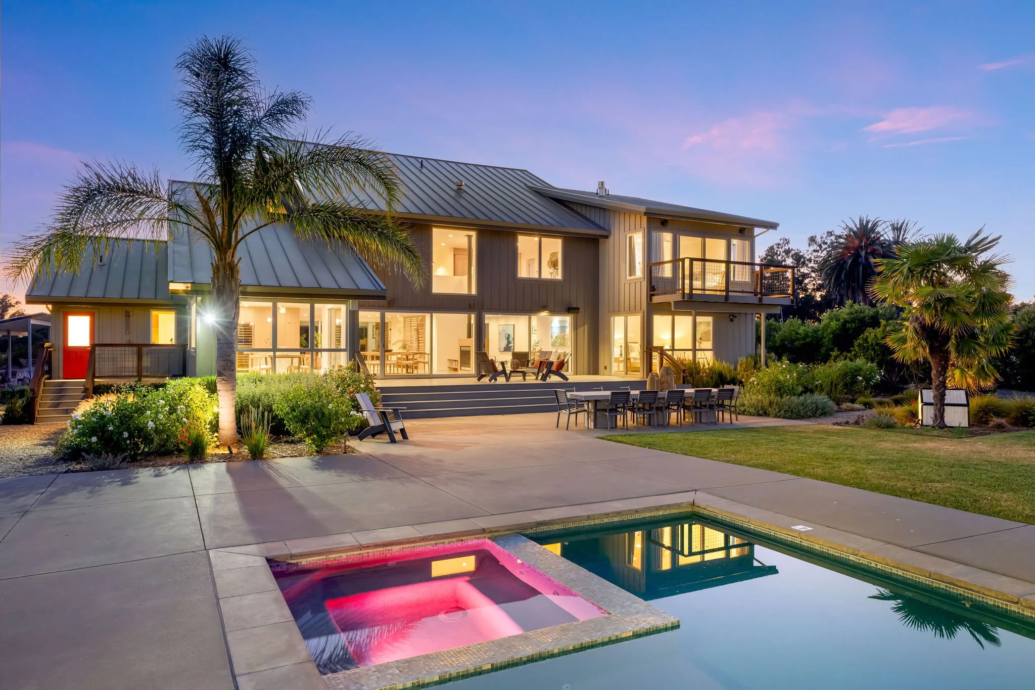 A modern two-story house with large windows, a balcony, and a backyard with a swimming pool and hot tub. The house is illuminated at dusk with garden and palm trees surrounding the area.