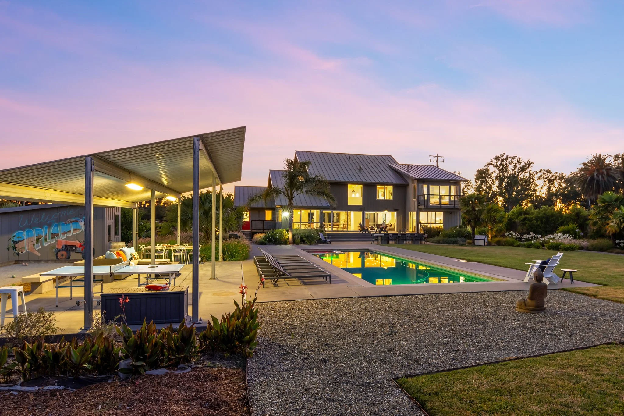 Luxury house with large windows, illuminated interior, backyard pool, outdoor seating area, palm trees, and landscaped garden during sunset.
