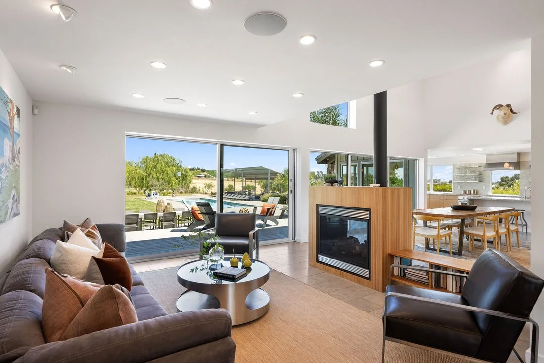 Modern living room with large sliding glass doors leading to an outdoor patio, a gray sofa with multiple cushions, a black armchair, a round coffee table with decor, and a wooden fireplace divider. Open kitchen and dining area visible in bright, airy