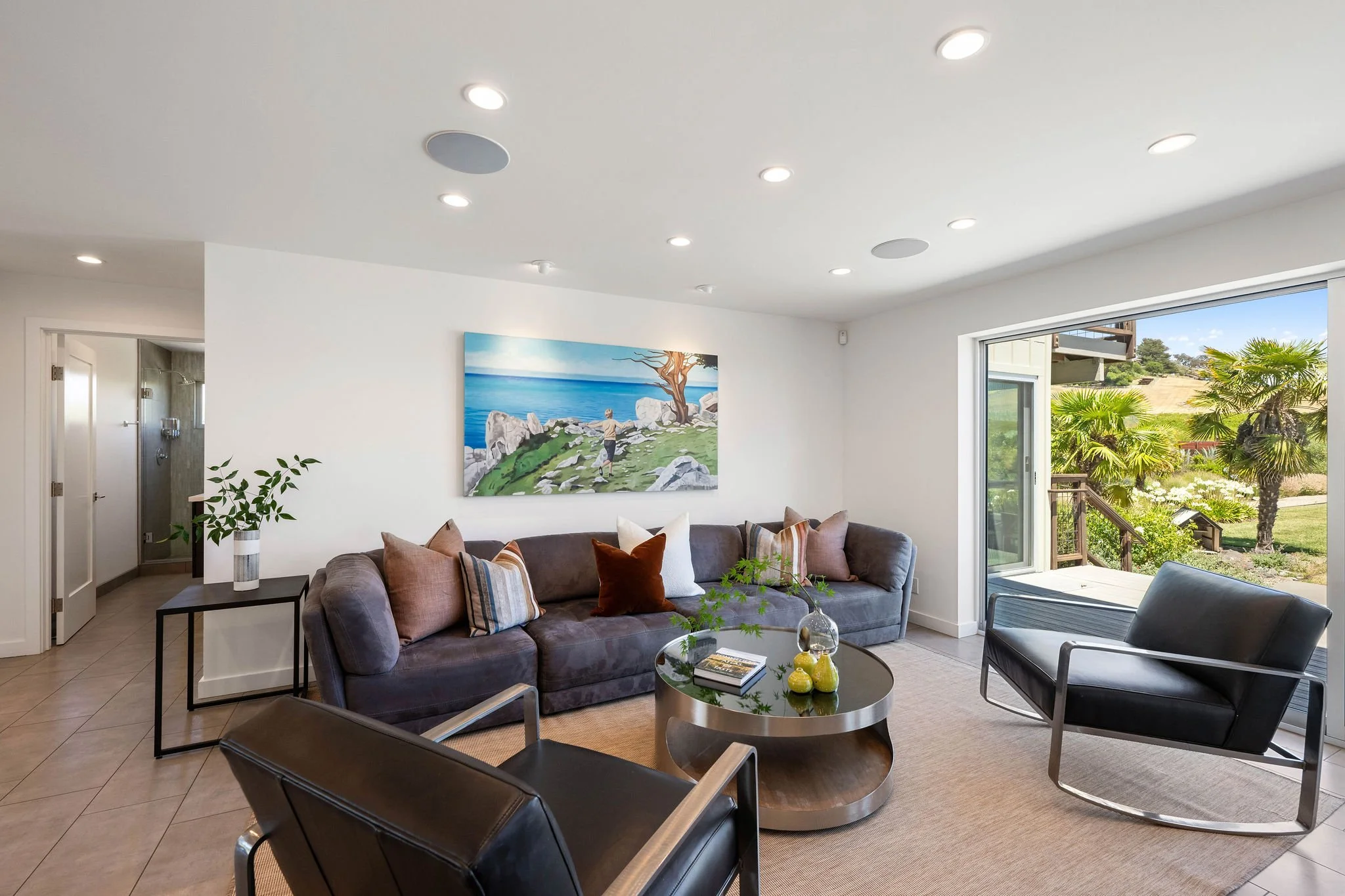 Living room with a gray sofa, black armchairs, and a round coffee table. There are decorative pillows on the sofa, a painting of a seaside scene on the wall, and large sliding glass doors opening to a garden with palm trees.