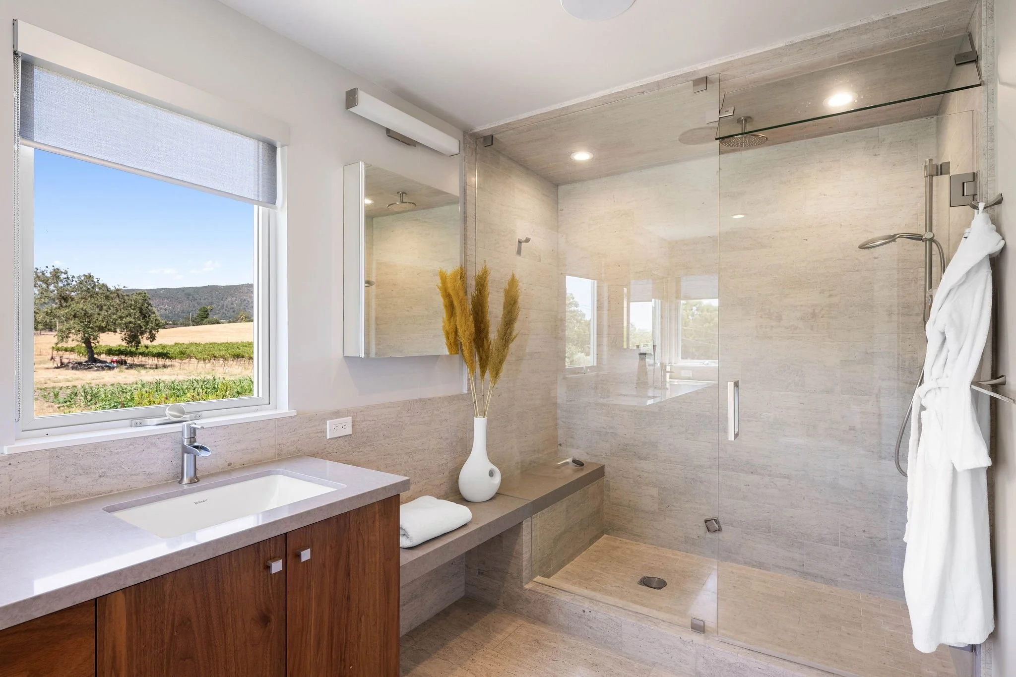 Modern bathroom with a large walk-in shower, a window showing a landscape, a wooden vanity with a white sink, a mirror, a white vase with dried plants, and a white robe hanging on a hook.