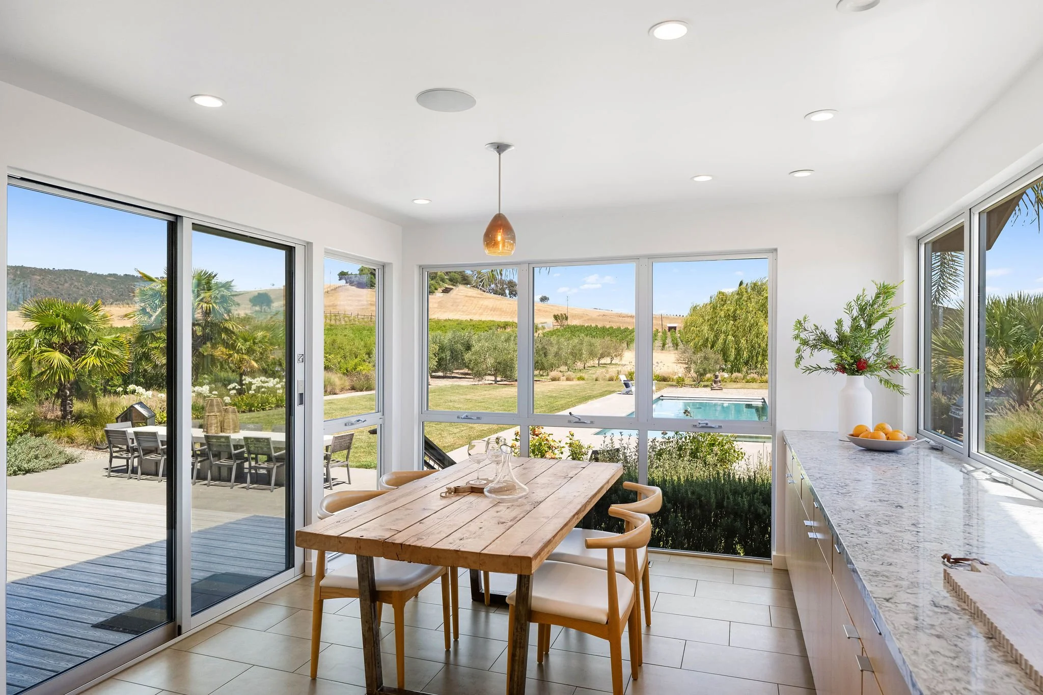 Bright dining area with a wooden table, six chairs, large windows showing backyard with patio, pool, trees, and hills, and a marble countertop with a vase of greenery and oranges.