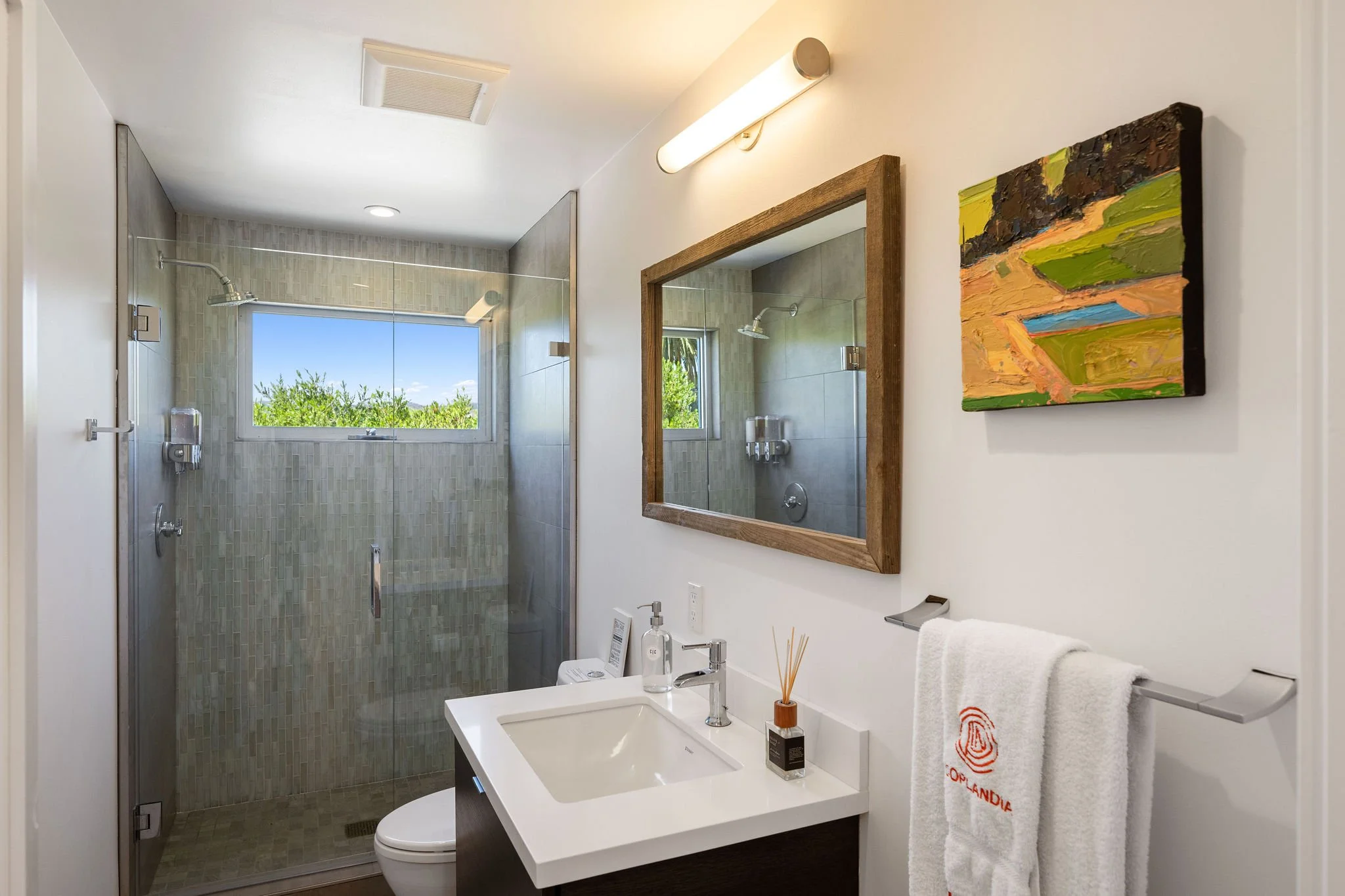 Modern bathroom with walk-in shower, glass door, window with greenery view, white sink, wooden framed mirror, abstract colorful painting, white towel with orange logo, soap dispenser, reed diffuser.