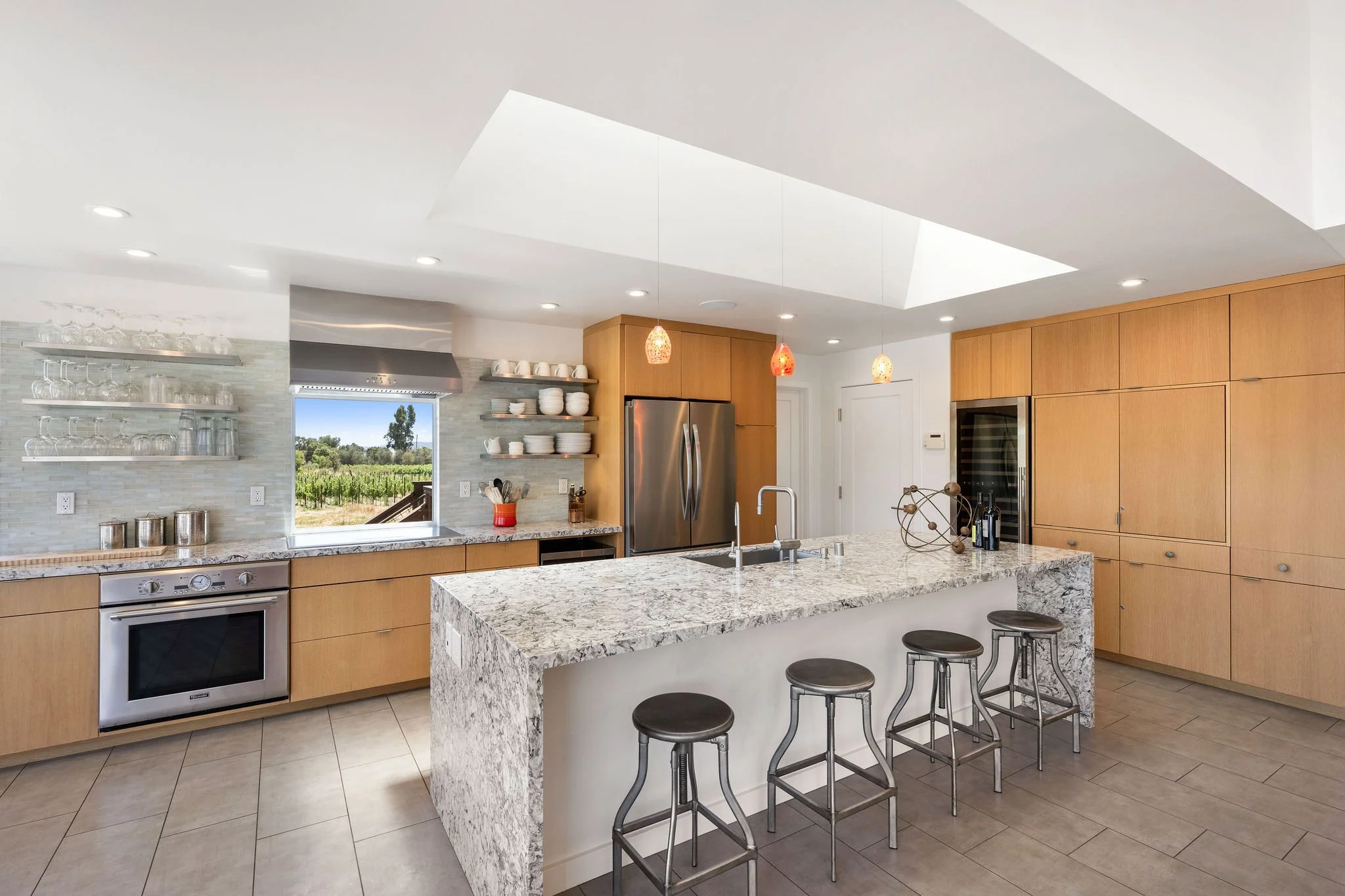Modern kitchen with wooden cabinetry, granite island, stainless steel appliances, open shelving with dishes and glasses, and a view of green fields through a window.