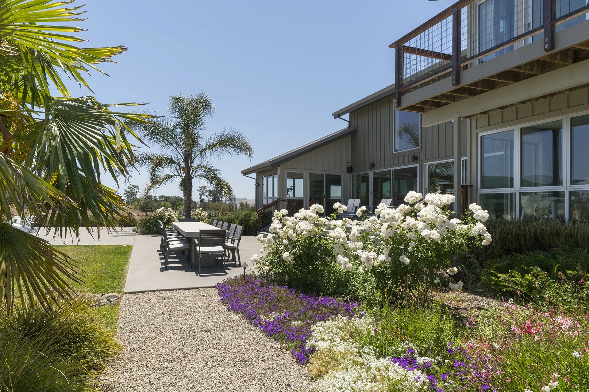 A backyard patio with a large table and chairs, surrounded by colorful flowers and palm trees, adjacent to a modern house with large windows and a balcony, under a clear blue sky.