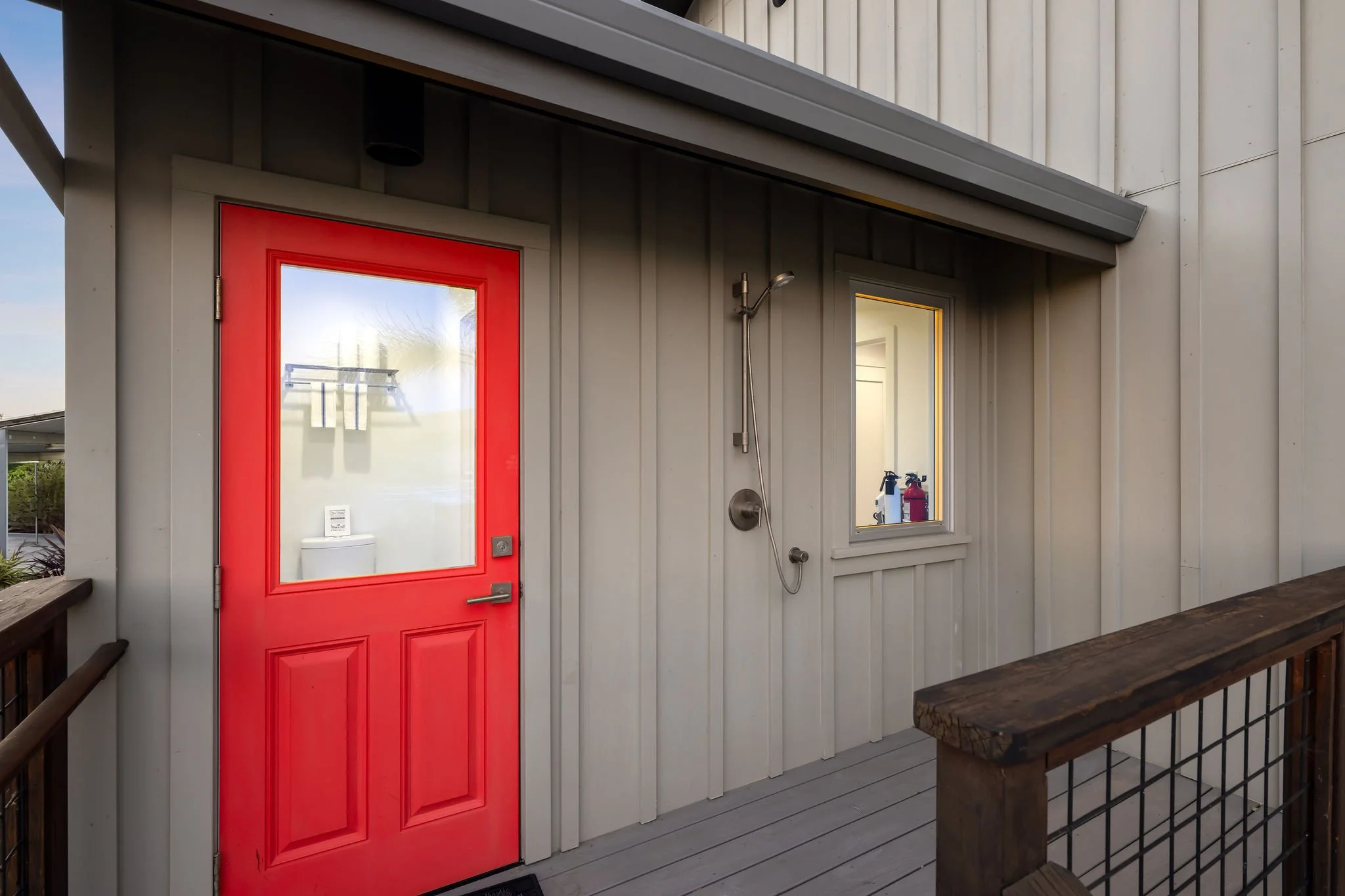 Exterior of a building with a red door with a large glass panel, an outdoor shower with a hose, and a window showing cleaning supplies inside.