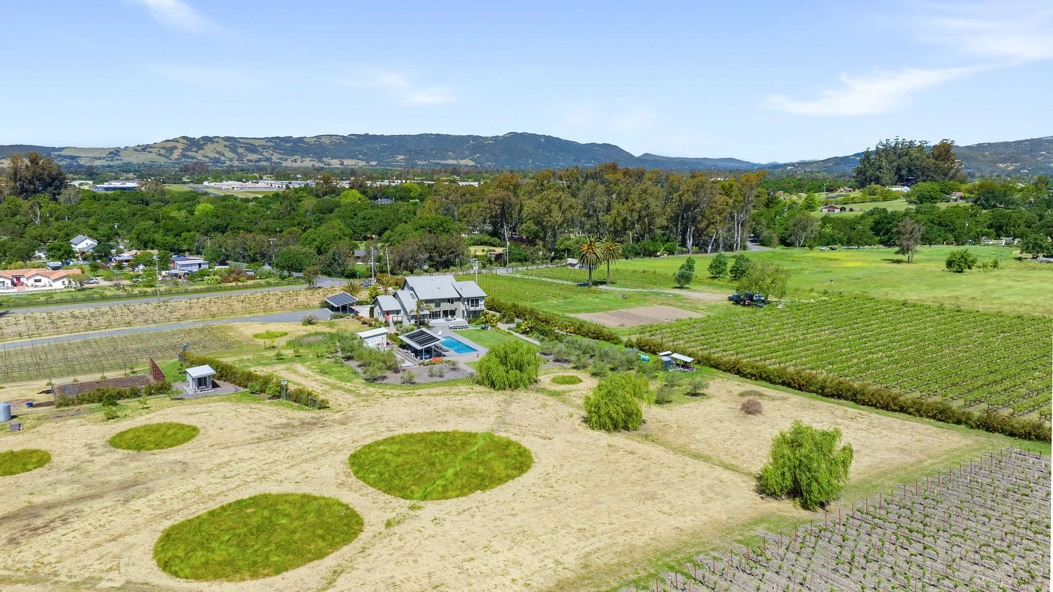 Aerial view of a rural landscape with farmland, vineyards, a house with a swimming pool, and surrounding green hills under a clear blue sky.