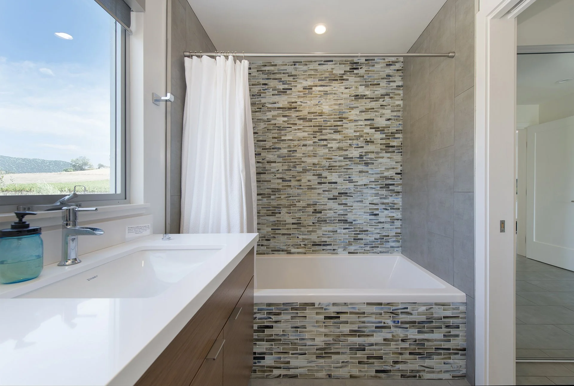 Modern bathroom with a window, white countertop with a sink, a blue soap dispenser, a bathtub with a mosaic tile wall backdrop, and a white shower curtain, beige tiled walls, and a doorway leading to a room with gray flooring.