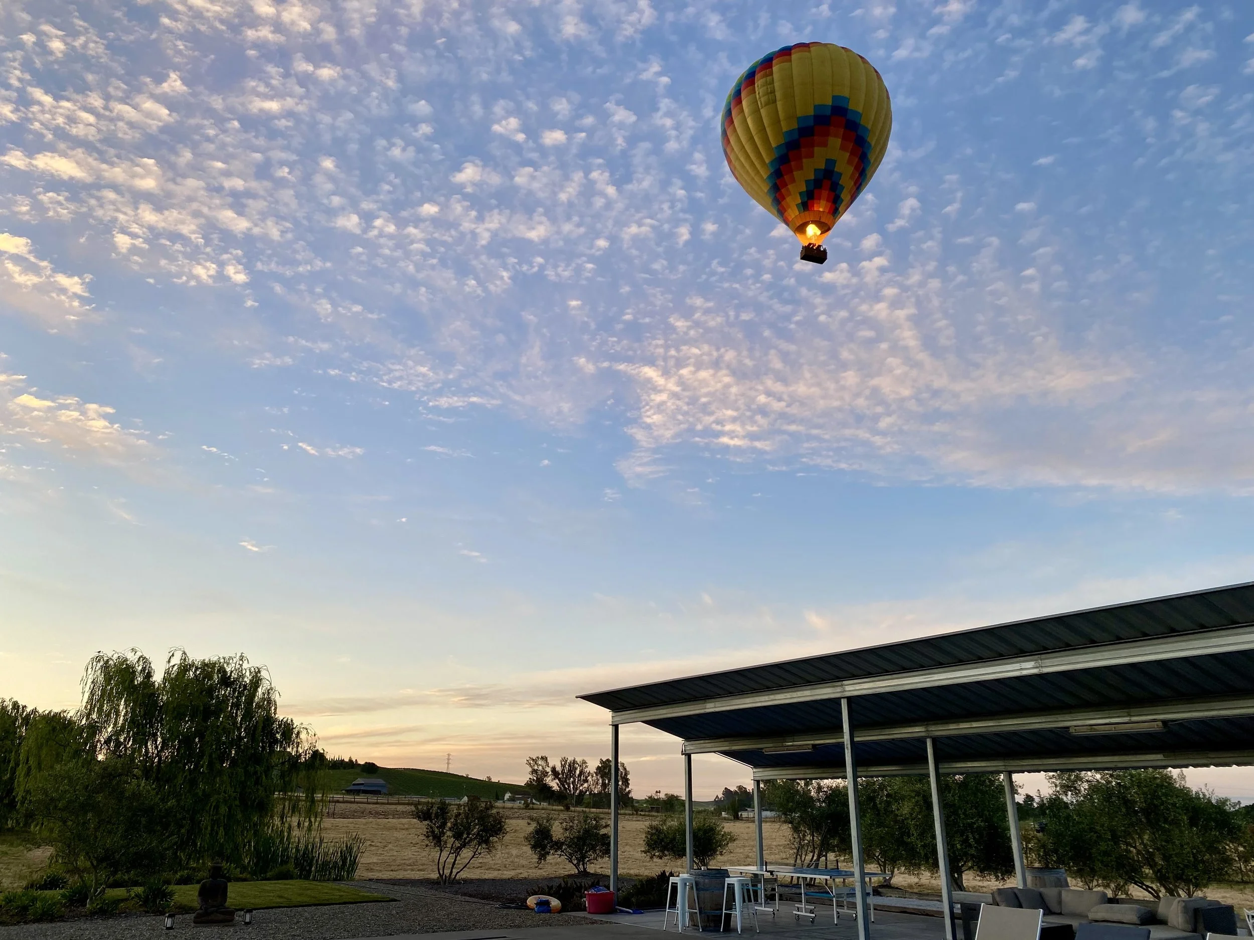 Colorful hot air balloon floating in a partly cloudy sky at sunset over a rural landscape with trees, a metal-roofed patio, and outdoor furniture.