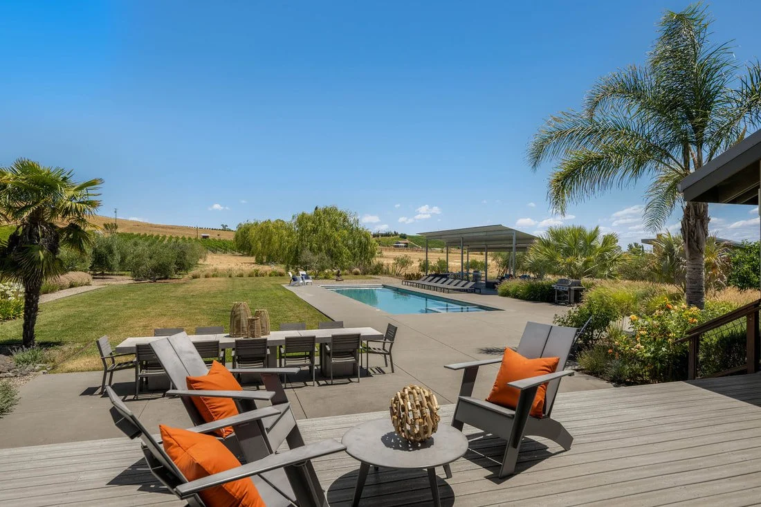 A backyard with a swimming pool, outdoor seating area with chairs and a table with orange cushions, a covered patio, and lush greenery including palm trees and bushes under a clear blue sky.
