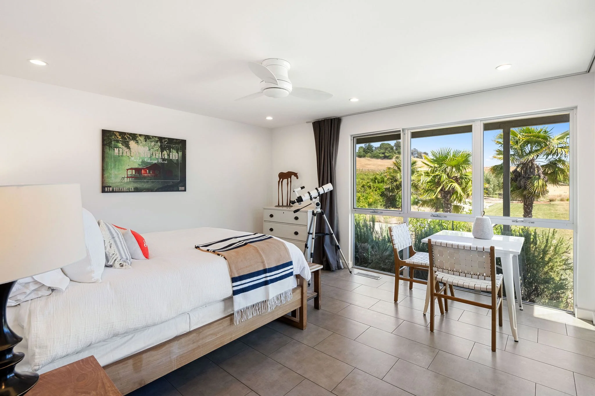 Bright bedroom with large window view of palm trees and greenery, white bed with pillows, striped blanket, telescope near dresser, small white table with two chairs.