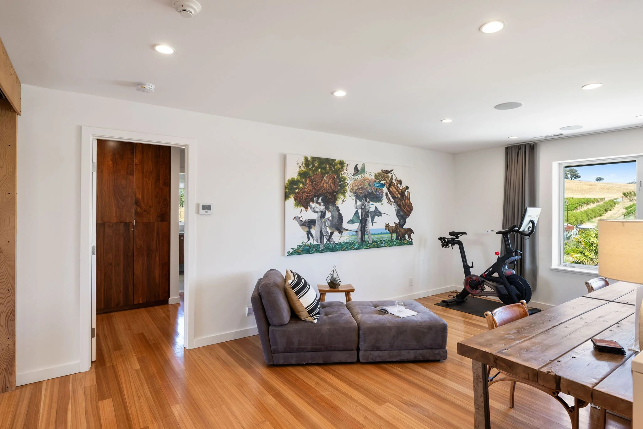 Living room with hardwood floors, white walls, large window, treadmill, couch with pillows, artwork on the wall, wooden dining table, and a door leading to another room.