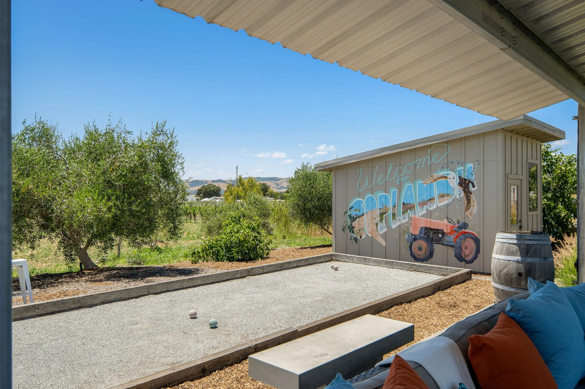 Officially titled "Welcome to Yollanda," the image depicts a plush outdoor bocce court with a painted mural on a beige shed reading "Welcome to Yollanda," featuring illustrations of a tractor, a bird, and landscape elements, set against a backdrop of