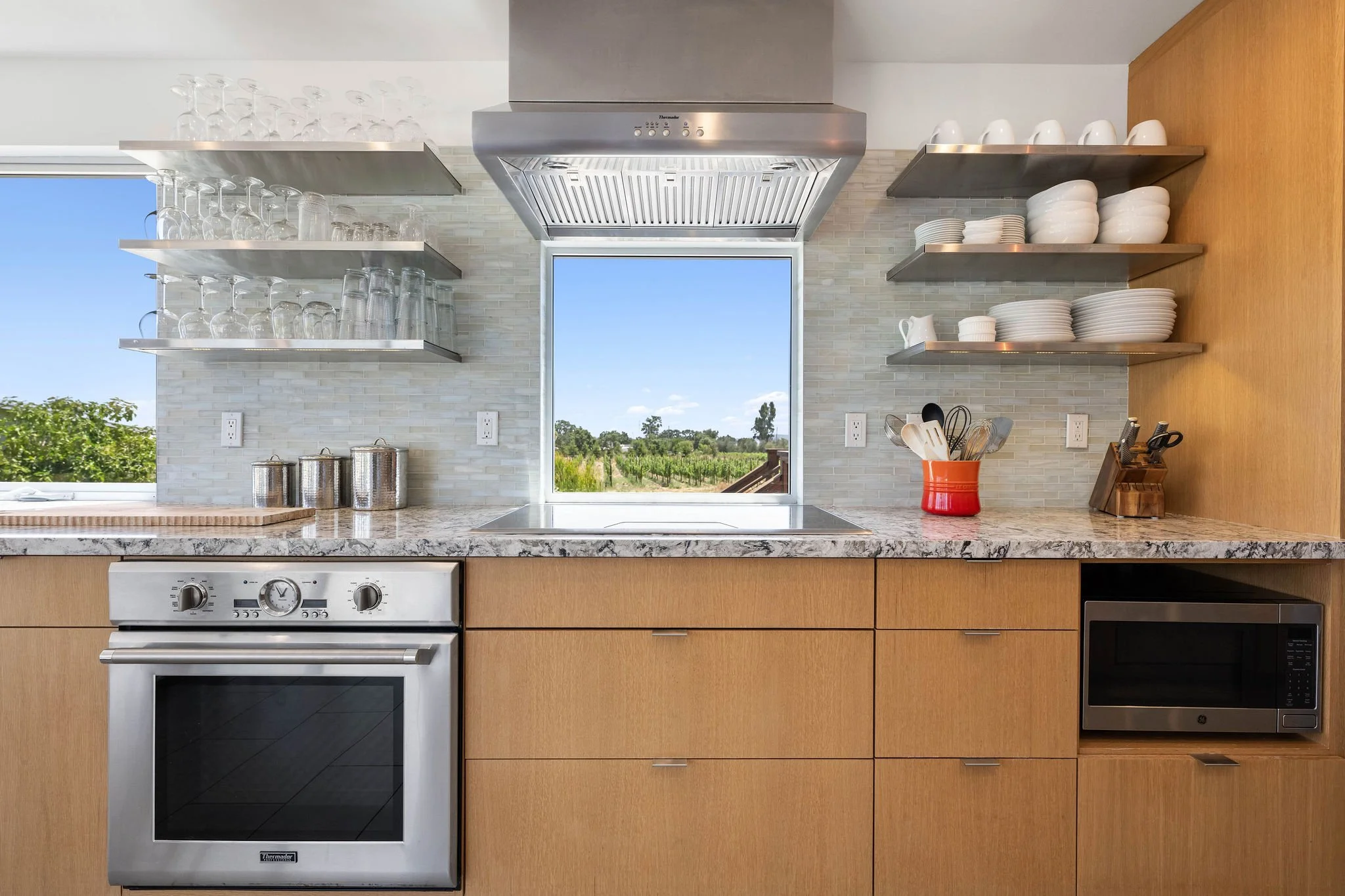 Modern kitchen with wooden cabinets, marble countertop, open shelves with white dishes and glasses, stainless steel oven, microwave, and vent hood, overlooking a window with a scenic outdoor view.