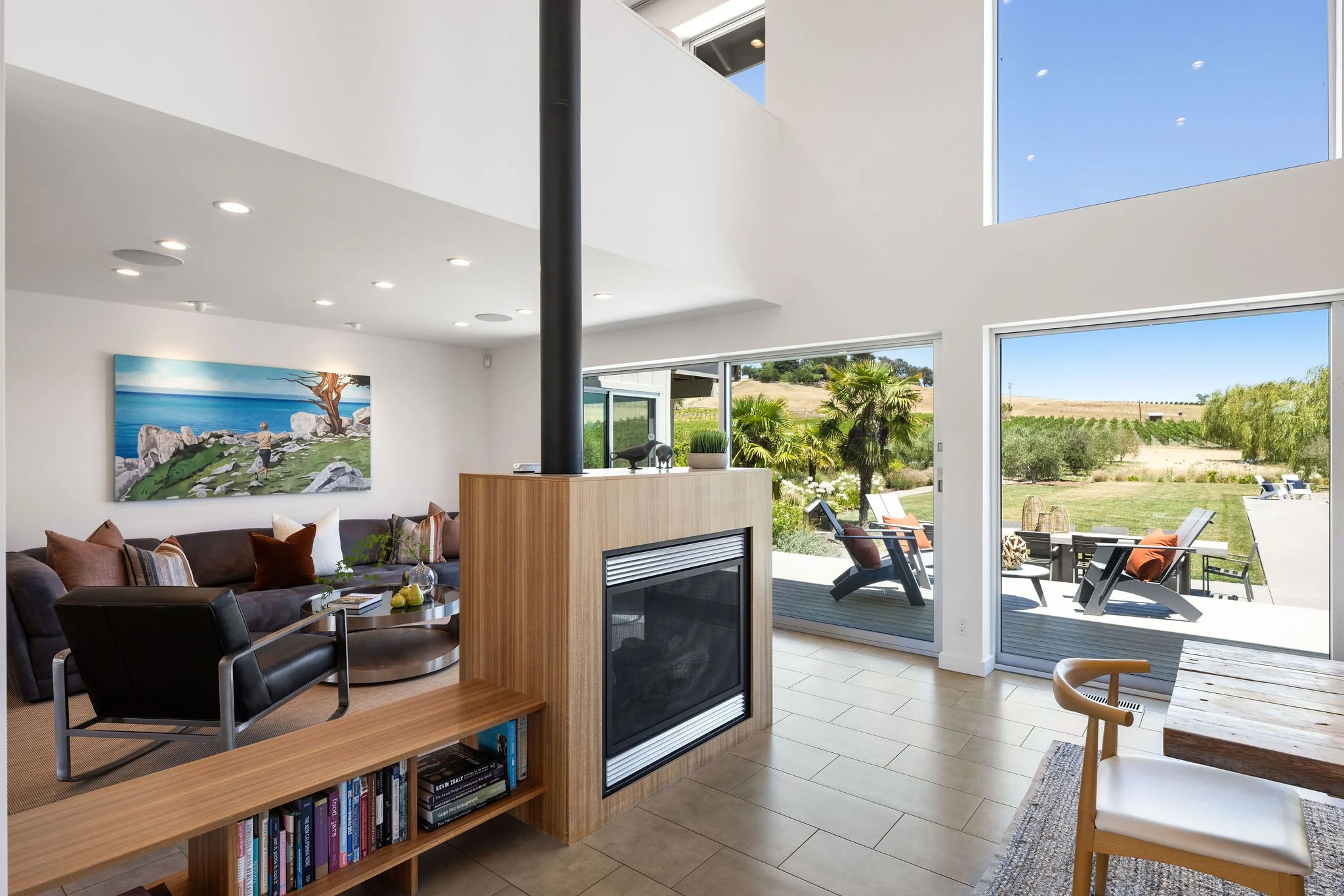 Modern living room with large glass sliding doors leading to outdoor patio; interior features a fireplace, bookshelf, black leather chair, brown sofa, and colorful cushions; artwork on wall depicts a seaside scene; outdoor patio has lounge chairs and