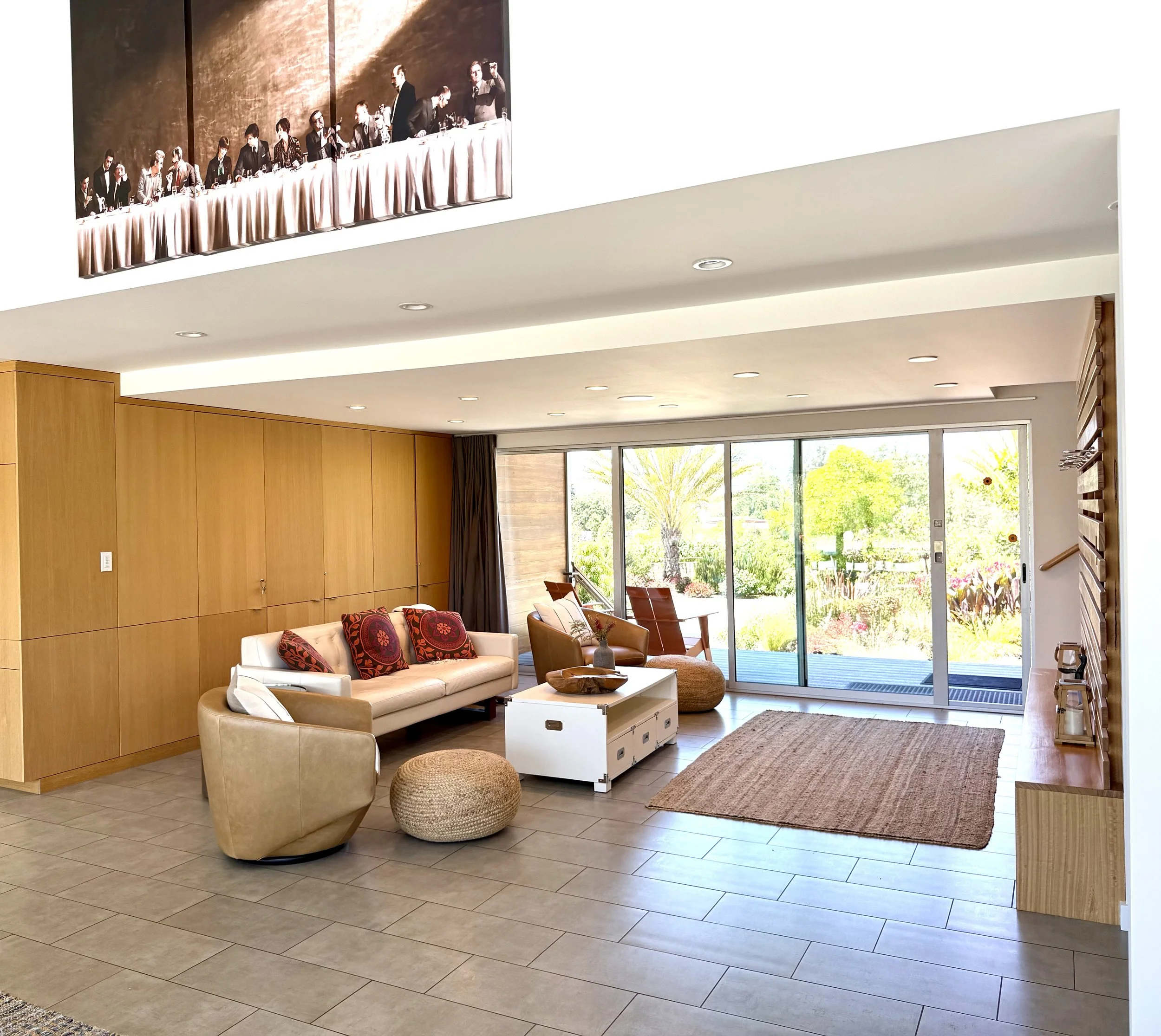 Living room with wooden wall panels, beige and white sofas, colorful pillows, a coffee table, a rug, and large sliding glass doors opening to a garden.