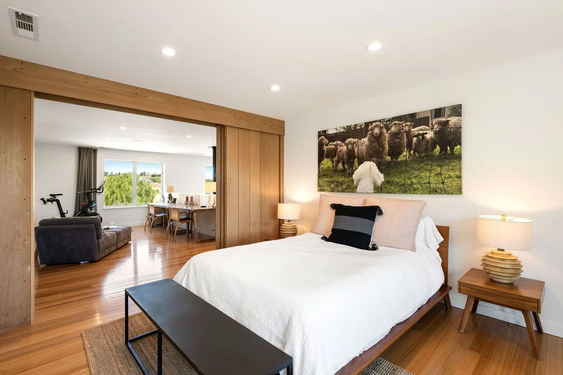 Bedroom with white bed, black pillow, wooden nightstands, lamps, large wall art of sheep, and a view into a living area with bicycle and dining table.