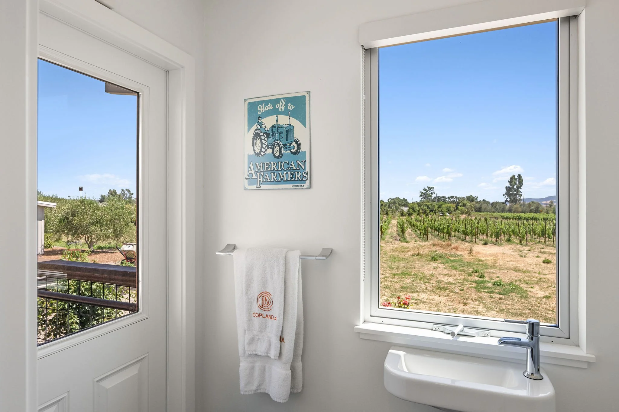 The image depicts a bright room with two large windows providing a view of a vineyard and clear blue sky. There's a white wall with a decorative poster of a tractor and the words 'Plant off to American Farmers.' A white towel is hanging on a metal to