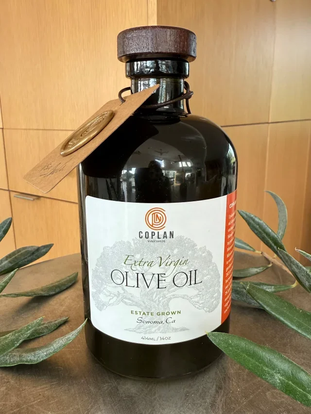 A bottle of Coplan Vineyards Extra Virgin Olive Oil, featuring a wooden cap and a label indicating it is estate-grown in Sonoma, California, surrounded by green leaves on a wooden surface.