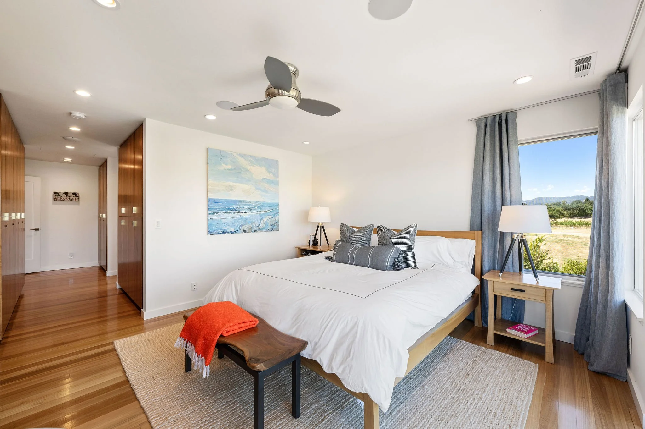 Bright bedroom with white walls, wooden floor, and a large window with blue curtains showing an outdoor landscape. The bed has white bedding, multiple gray pillows, and a wooden headboard. There are two nightstands with lamps, and a painting of an oc