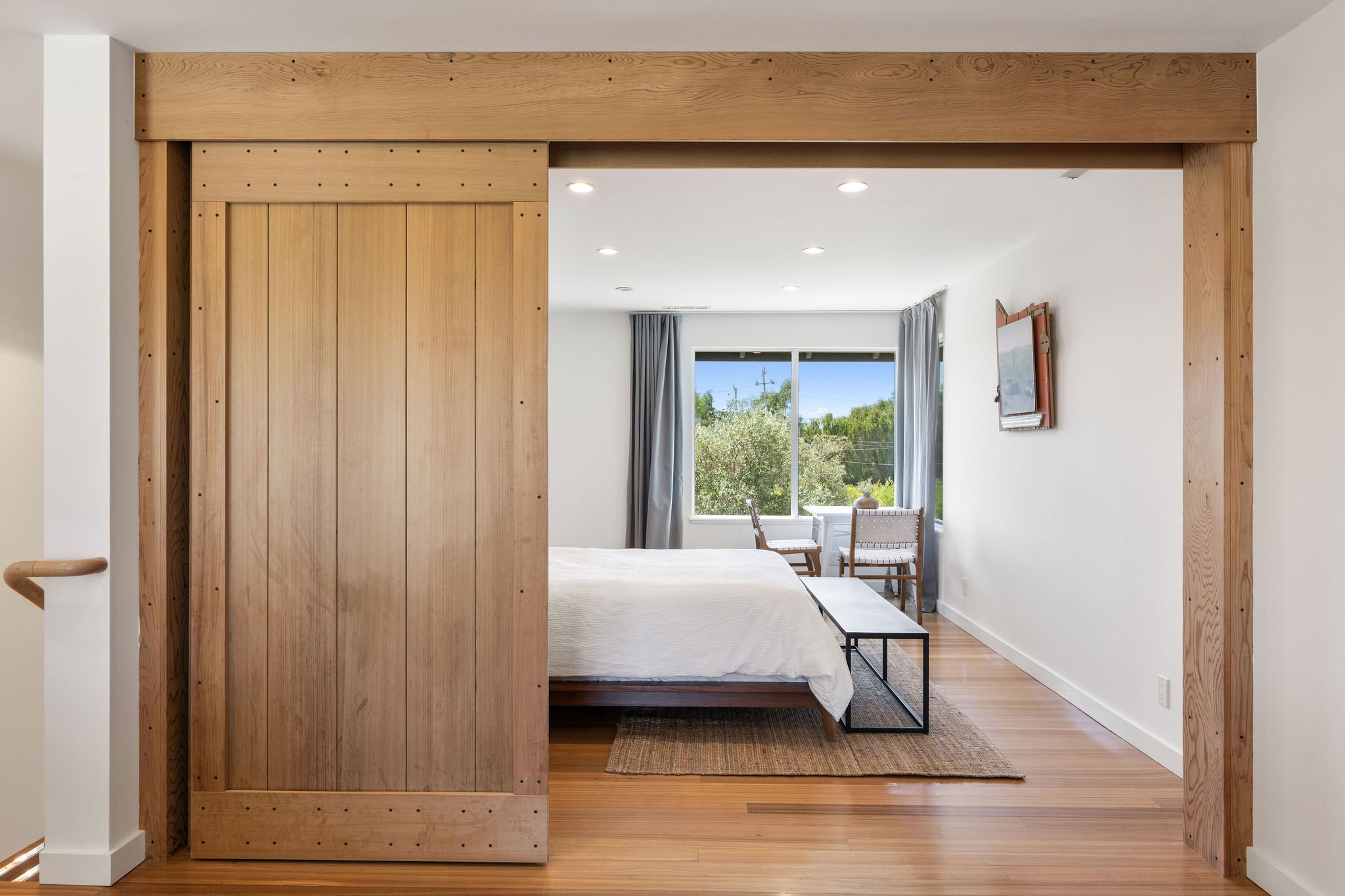 Bedroom with a sliding wooden door, white bed, black metal bench, window with gray curtains, and a small table with chairs