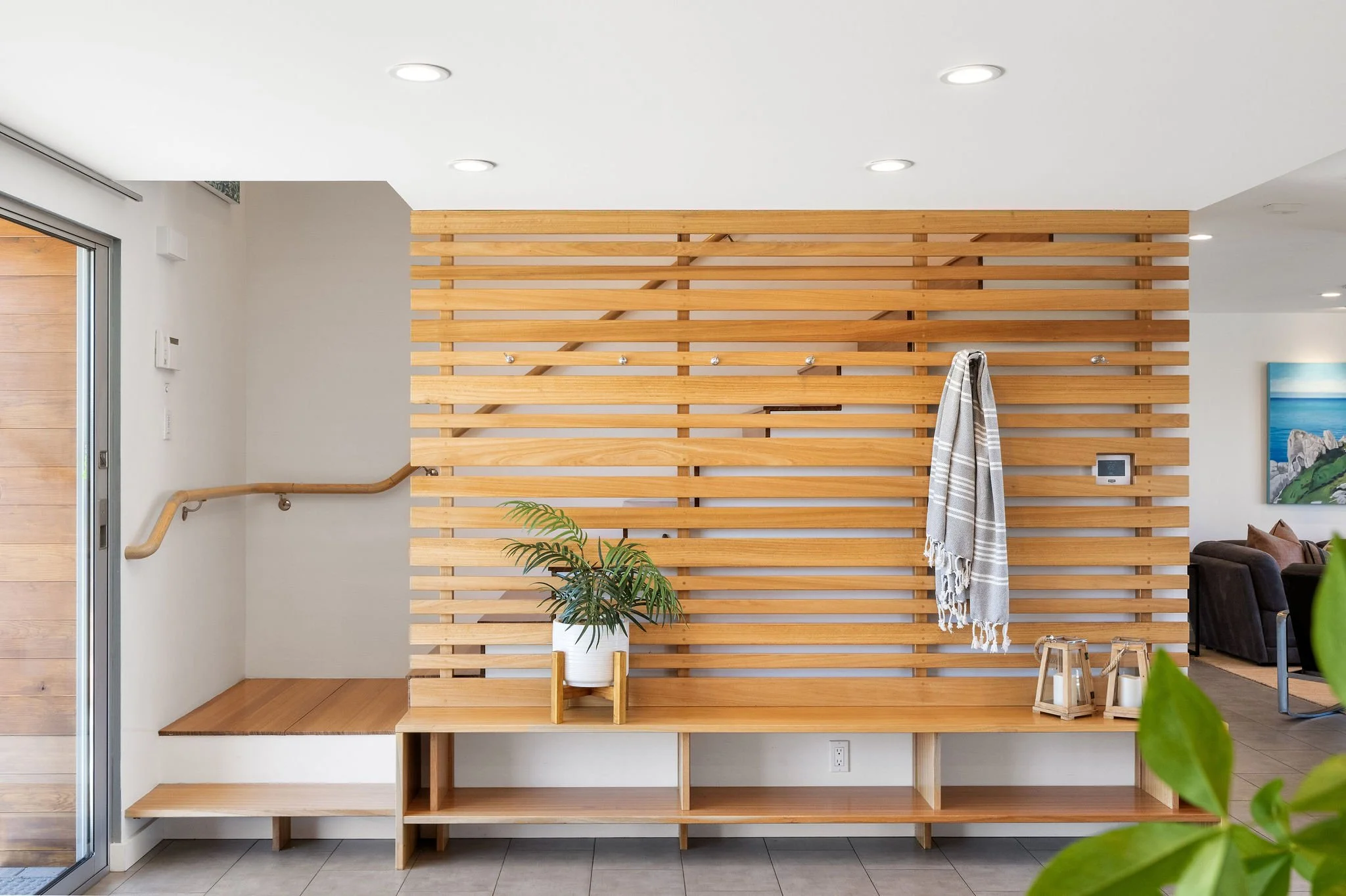 Interior view of a modern home with a wooden slat accent wall, a built-in bench, and a potted plant on top, with a staircase and living room visible in the background.