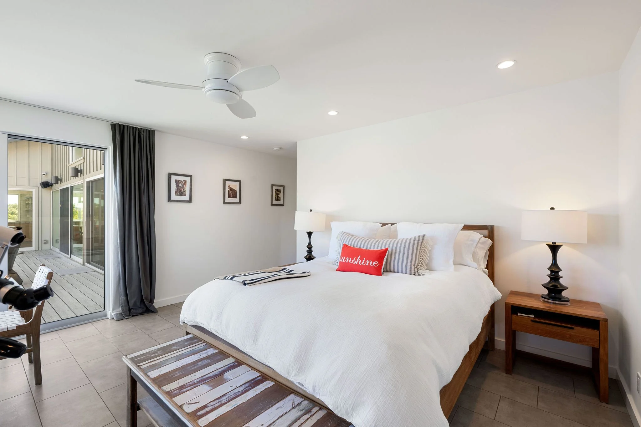 Bright, modern bedroom with king-sized bed, white bedding, decorative pillows, wooden nightstand, black table lamp, ceiling fan, sliding glass door leading to balcony, framed artwork, and neutral tile flooring.