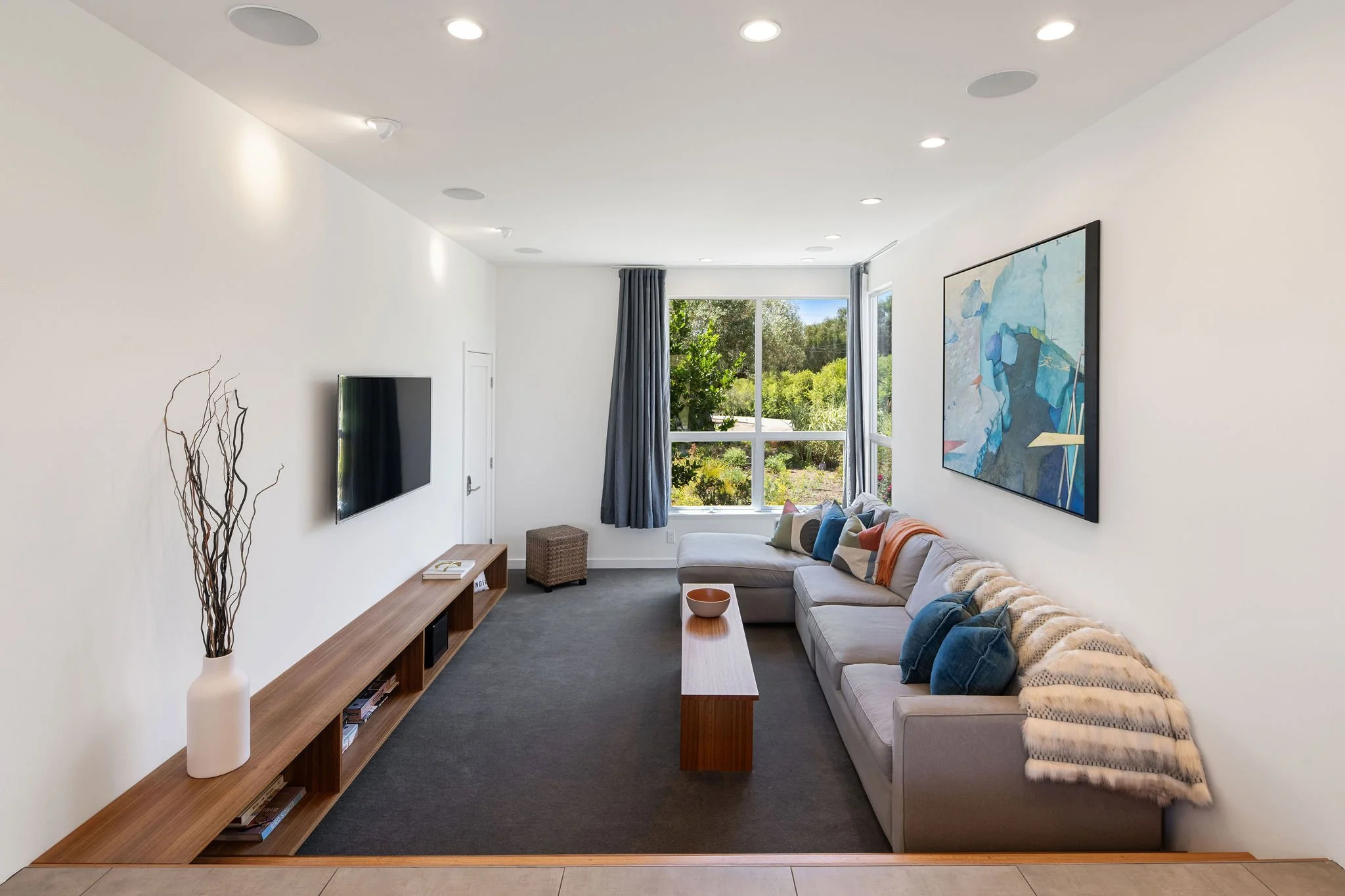 Bright living room with a large window, gray sofa with colorful pillows, a wooden coffee table, a wall-mounted TV, a modern painting, and a long wooden media console with decorative items and books.