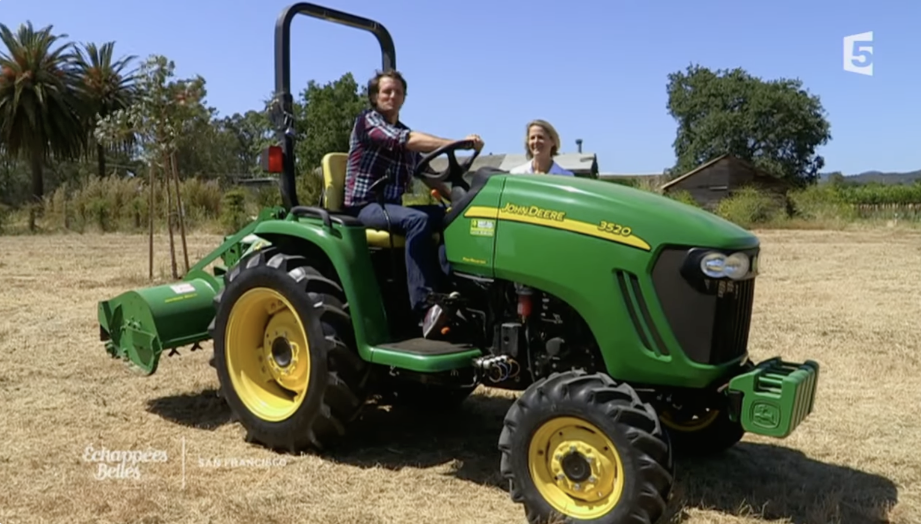Man and woman on a green John Deere tractor in a rural field with trees and a small building in the background.