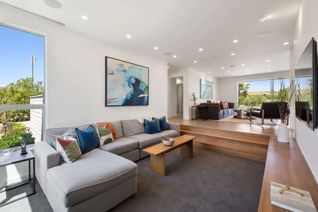 Modern living room with white walls, large windows, and wooden floors. There is a white sectional sofa with colorful pillows, a small black side table, and a wooden coffee table. In the background, there is a raised seating area with additional couch